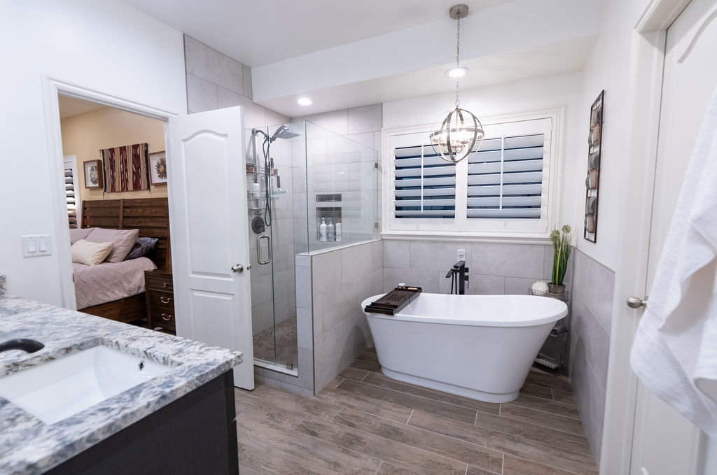 Bathroom featuring a freestanding soaking tub, glass-enclosed shower, wood-finish tile flooring, a window with plantation shutters, and a vanity with a white countertop and undermount sink