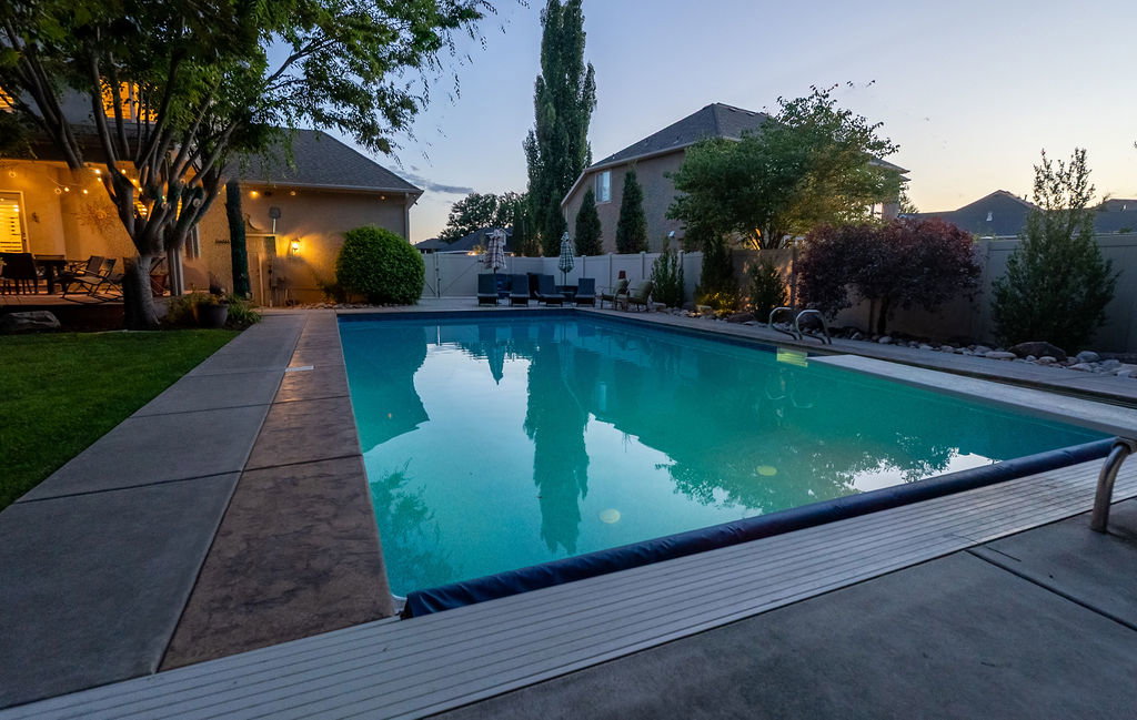 Outdoor swimming pool with concrete decking, surrounding green lawn, perimeter landscaping, and a covered patio with string lights