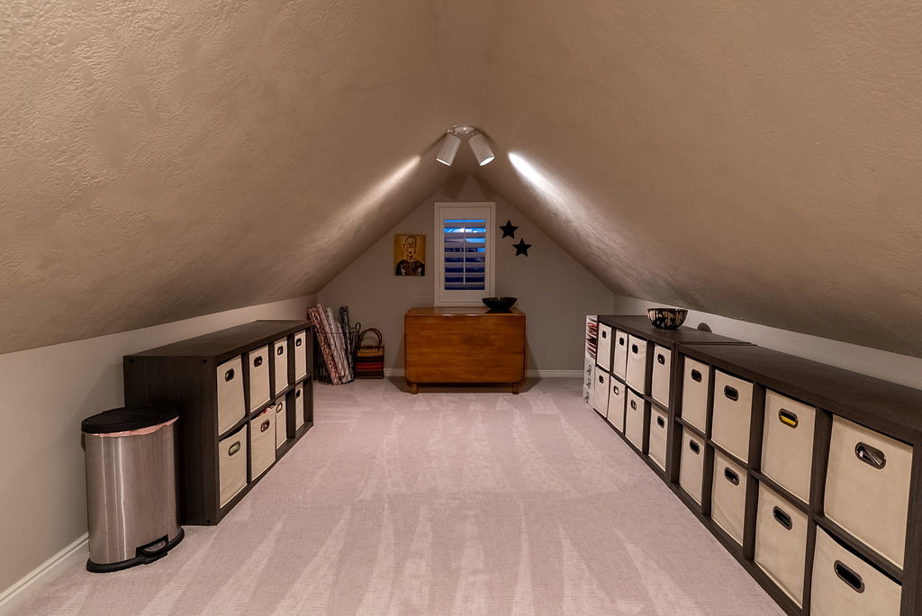 Finished attic space featuring sloped ceilings, light-colored carpeting, a window with plantation shutters, and track lighting