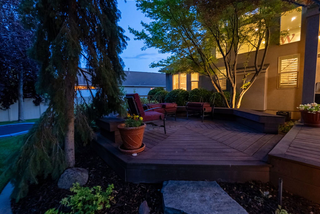 Expansive multi-level wood-finish deck featuring built-in bench seating, mature landscaping, and accent lighting
