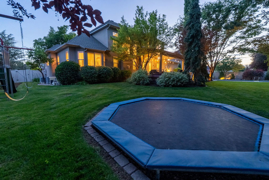 Expansive rear yard featuring a manicured lawn, established landscaping, and an in-ground trampoline