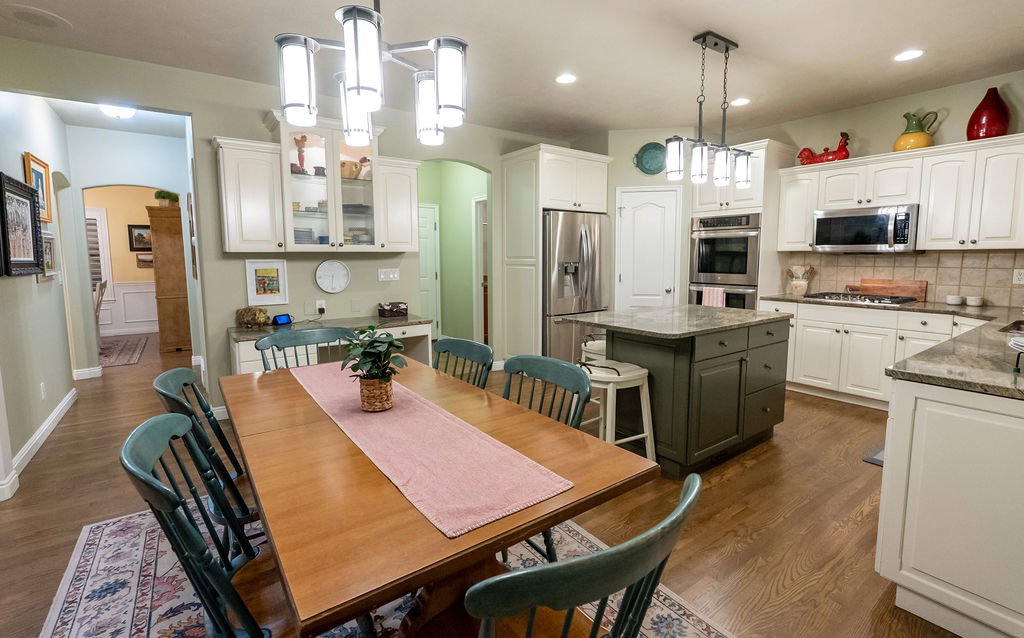Spacious kitchen and dining area featuring wood-finish flooring, white cabinetry, stainless steel appliances, a green kitchen island with a stone countertop, and a tiled backsplash