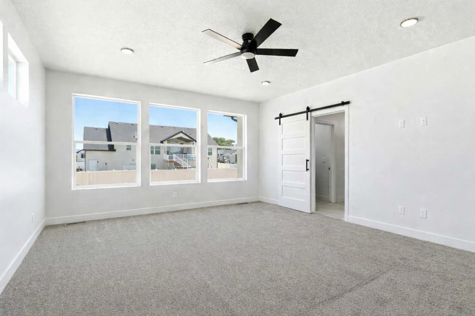 Spacious room featuring three large windows, light gray carpet, and a modern black ceiling fan