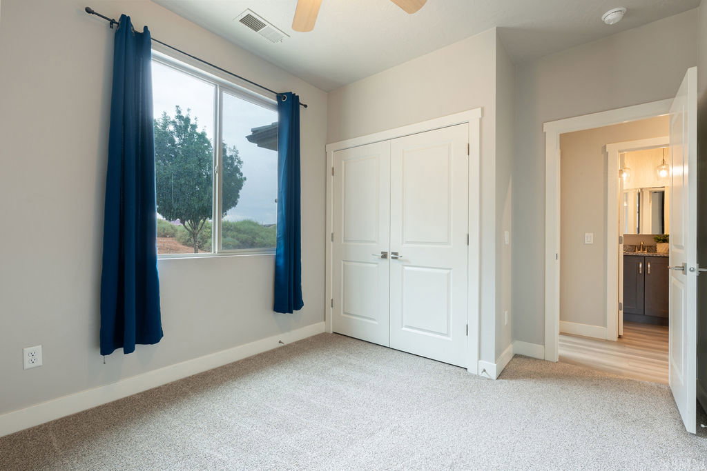 Carpeted room featuring a large window, white trim, a double-door closet, and an open doorway revealing a bathroom with wood-finish flooring and a vanity