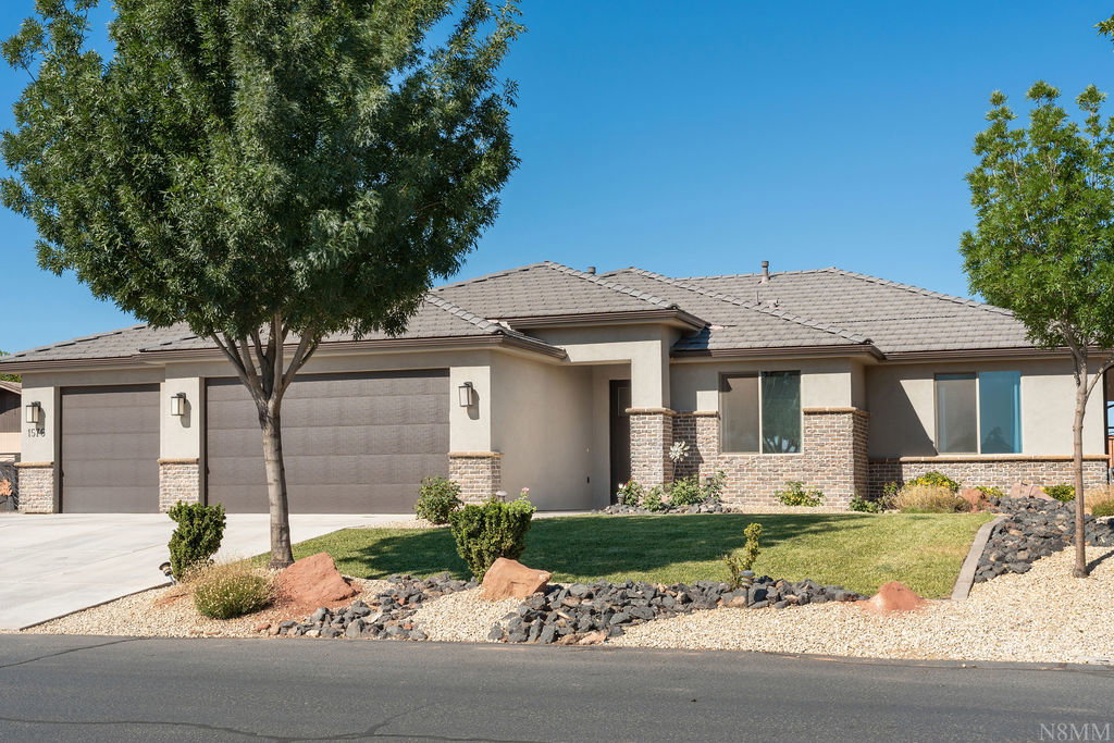 Ranch-style residence featuring a stucco and stone exterior, two-car garage, concrete driveway, tiled roof, and xeriscaped front yard with mature trees