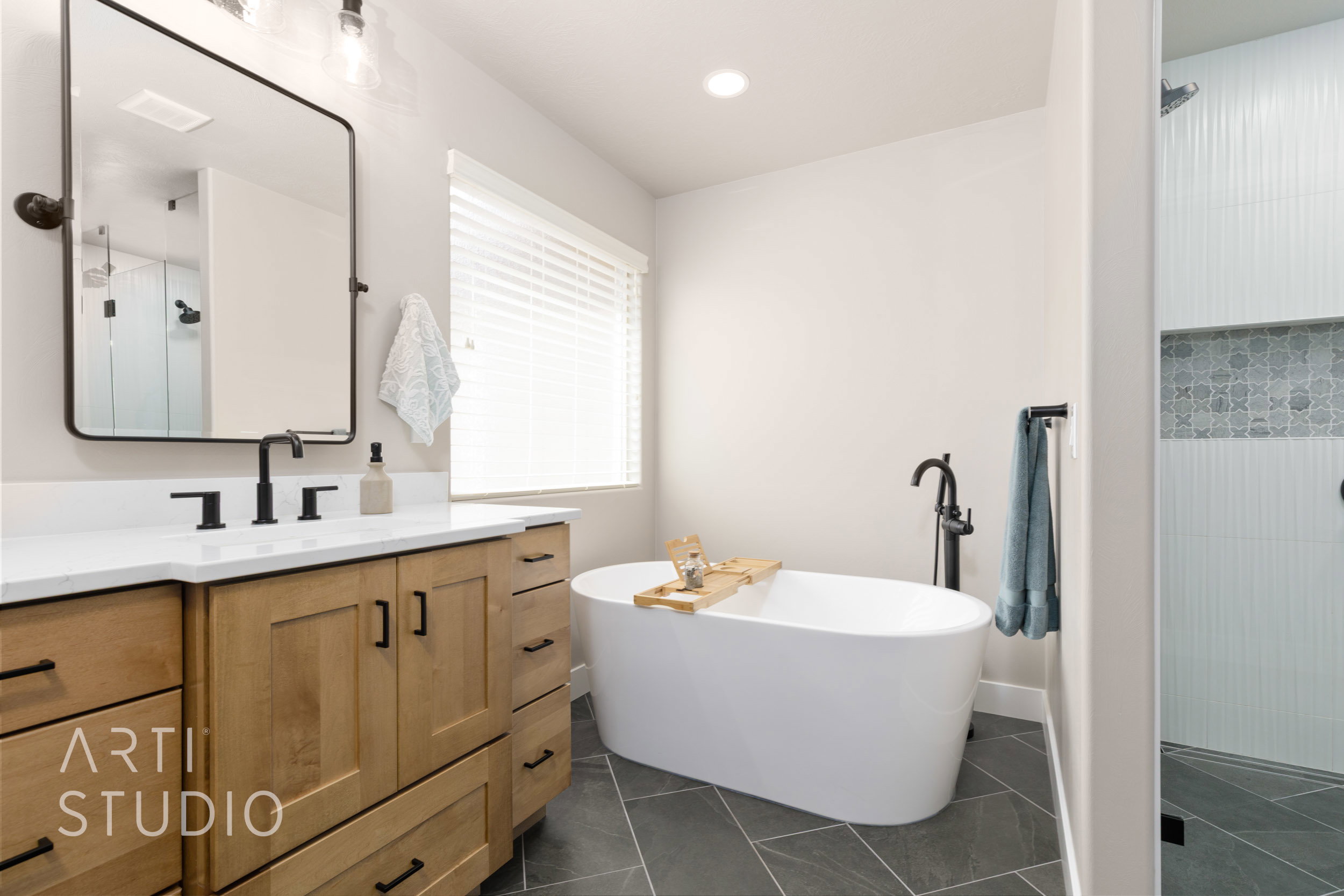 Bathroom featuring a freestanding soaking tub with a floor-mounted faucet, dual vanity with a white countertop and wood-finish cabinetry, a rectangular mirror with a black frame, and a shower with patterned accent tiling