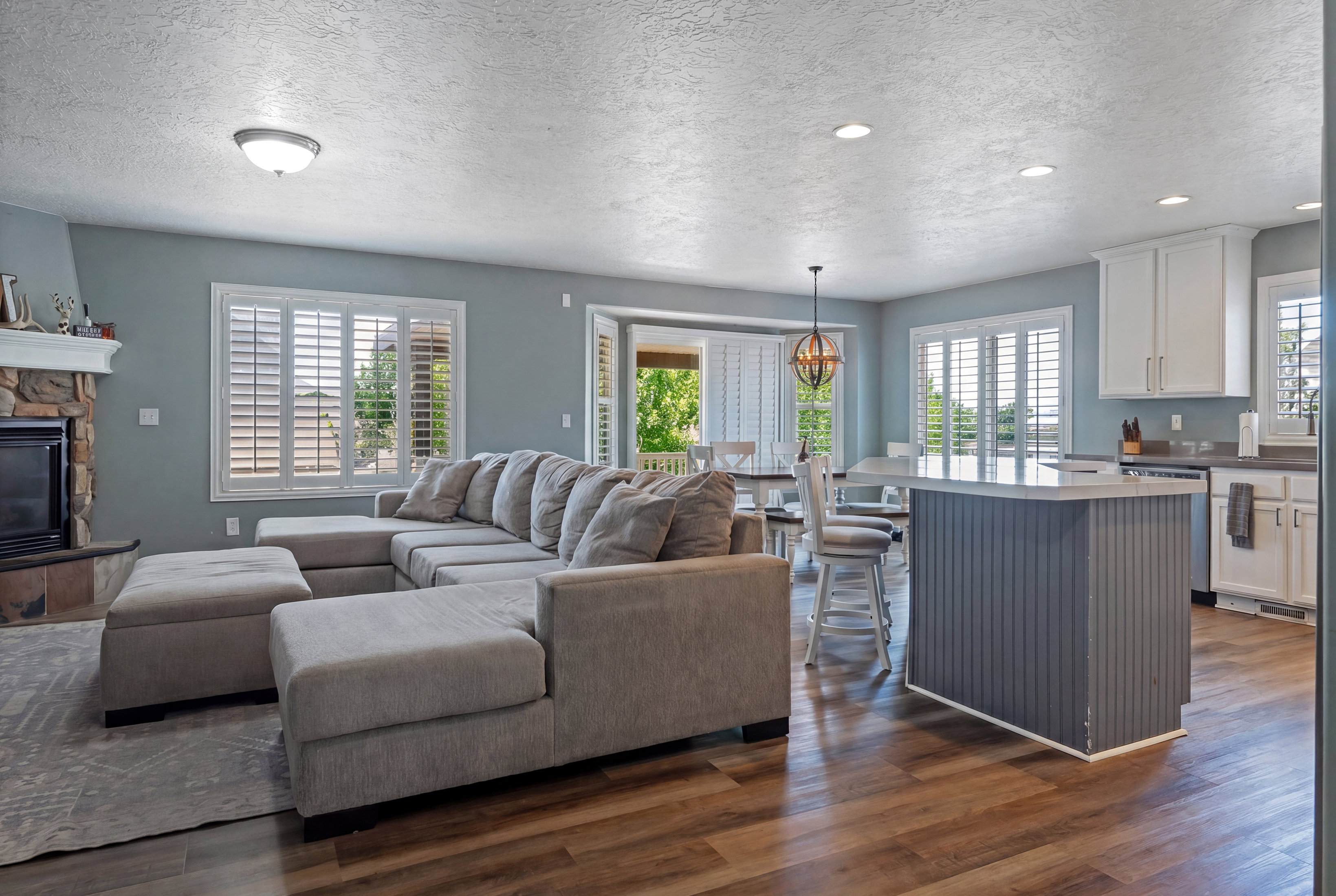 Open-concept living area with wood-finish flooring, a stone-faced fireplace, and white plantation shutters