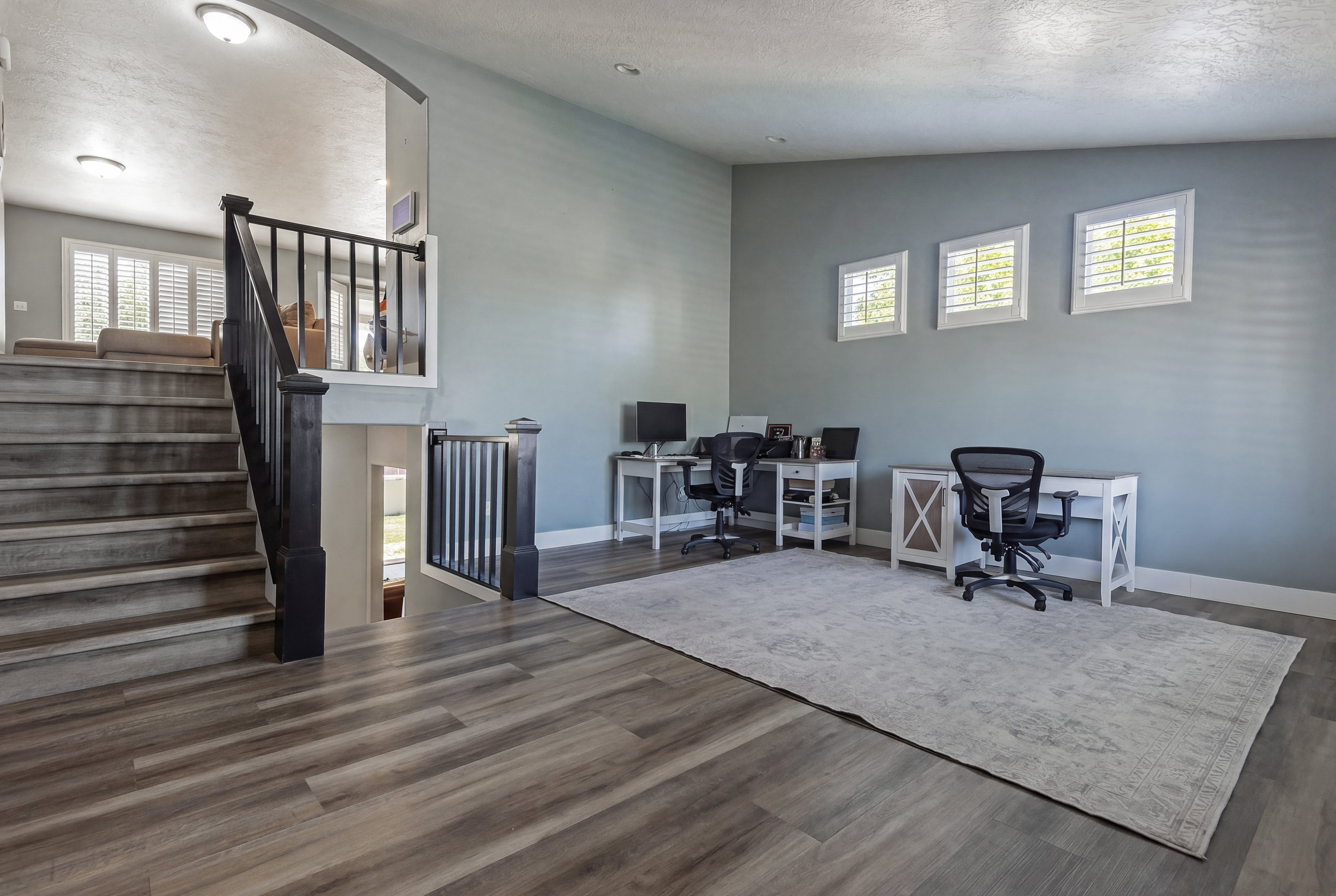 Spacious open-concept area featuring wood-finish flooring, a contemporary staircase with dark wood newel posts and spindles, three windows with plantation shutters, and light blue wall paint