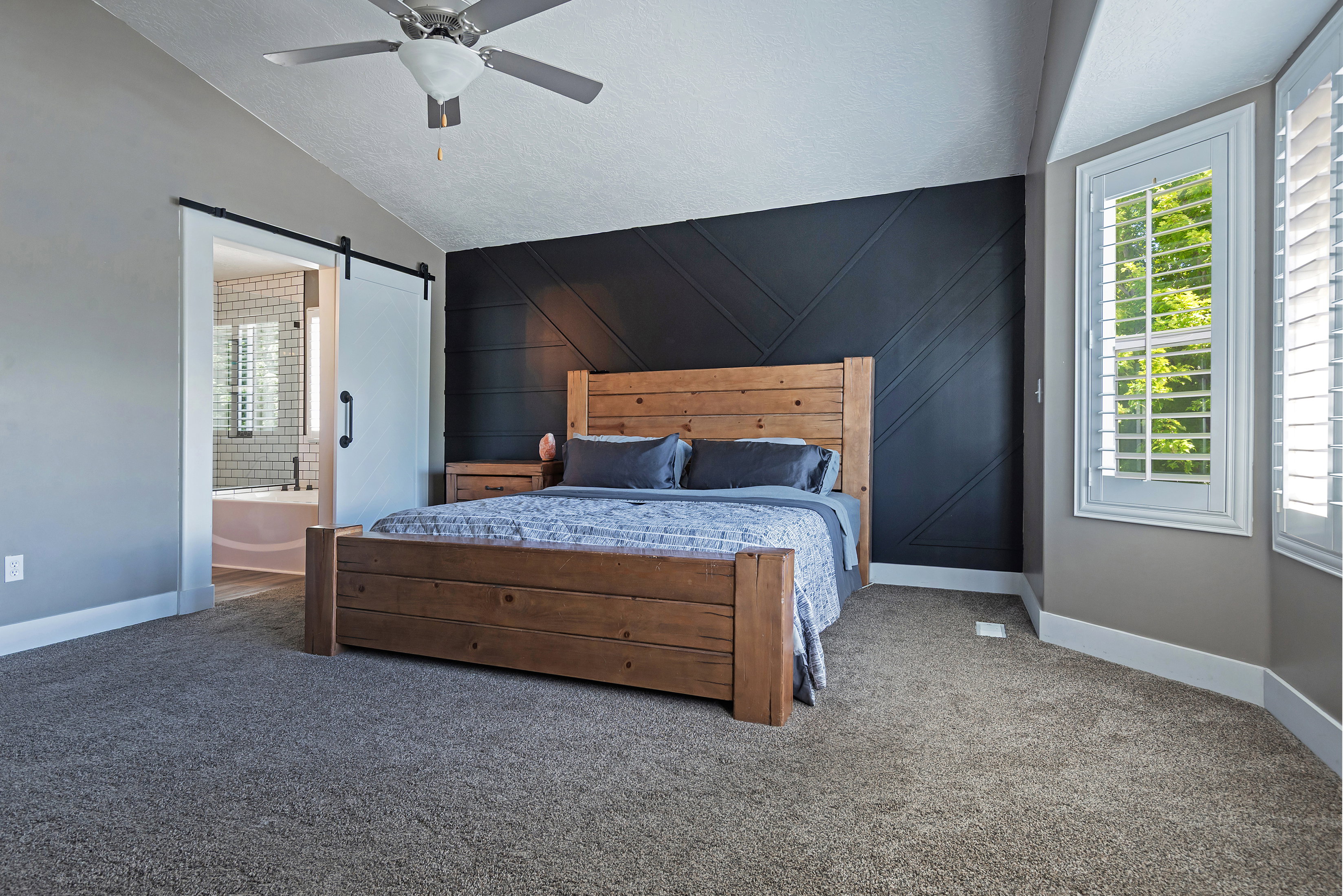 Vaulted ceiling with a ceiling fan, accent wall with geometric paneling, bay window with plantation shutters, barn door entry to a bathroom, and plush carpet flooring