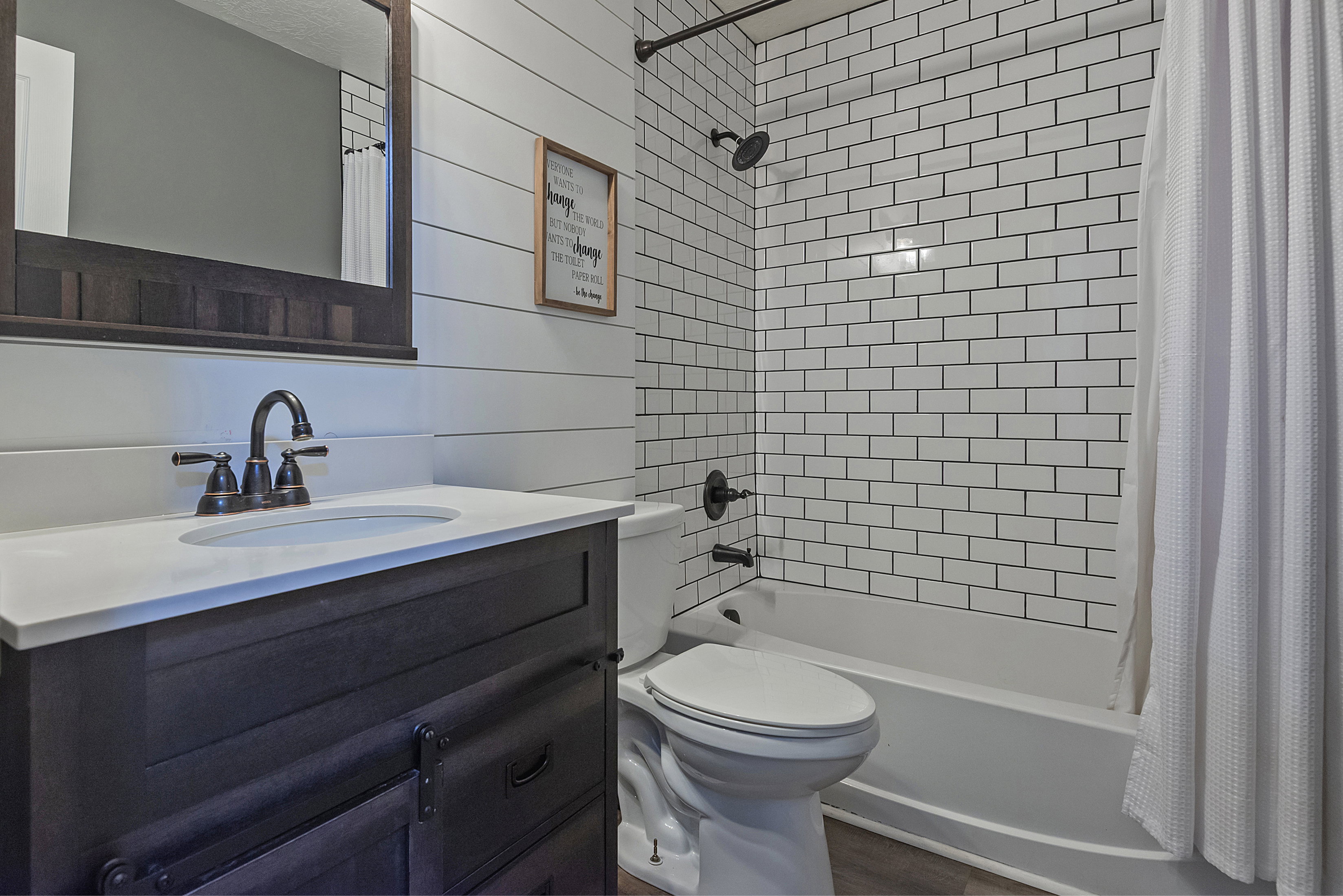 Bathroom featuring a dark wood-finish vanity with a white countertop and oval sink, dark bronze fixtures, white shiplap wall paneling, and subway tile shower surround
