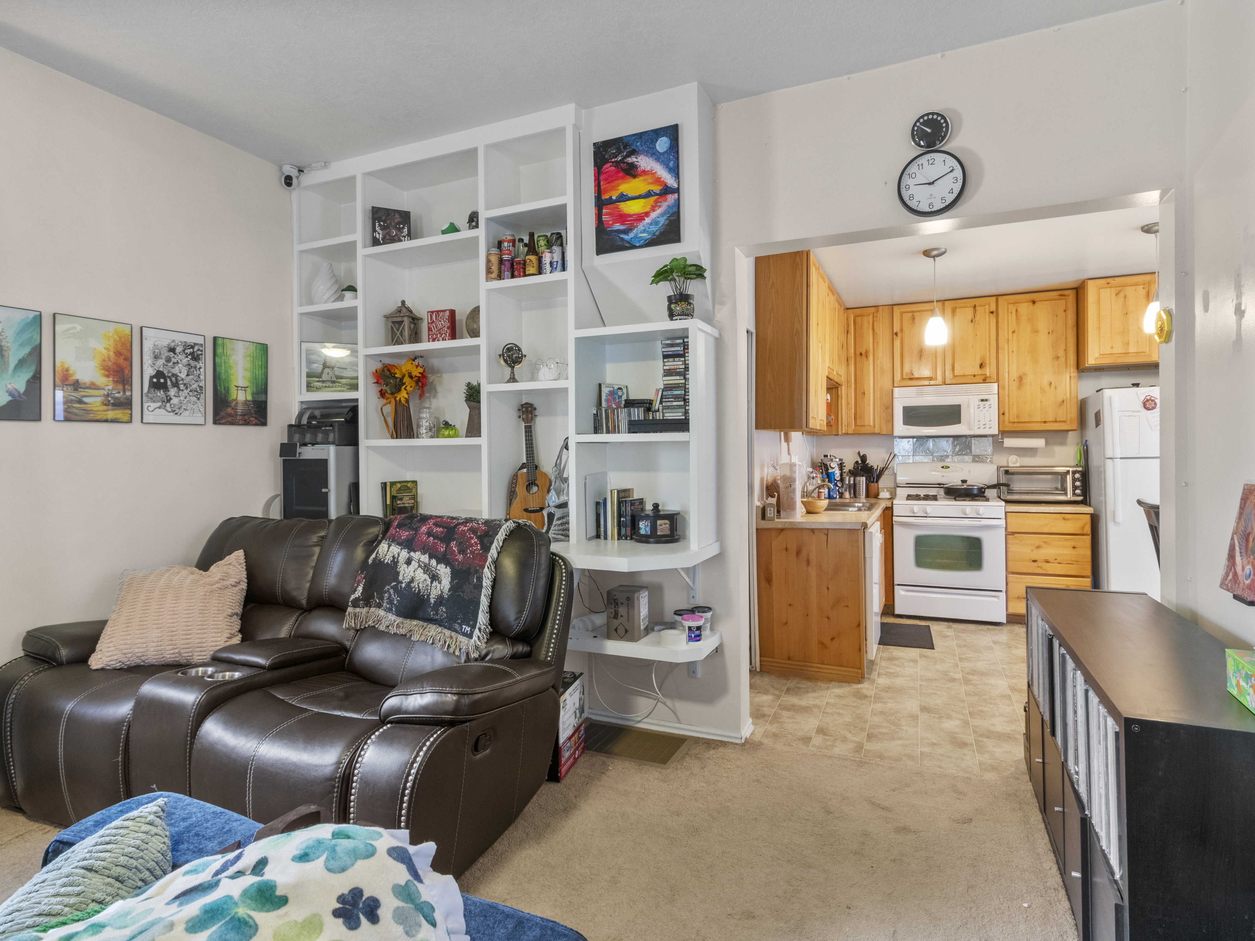 Built-in white shelving unit in a living area featuring an archway entrance to a kitchen with light wood cabinetry, white appliances, and a tiled backsplash