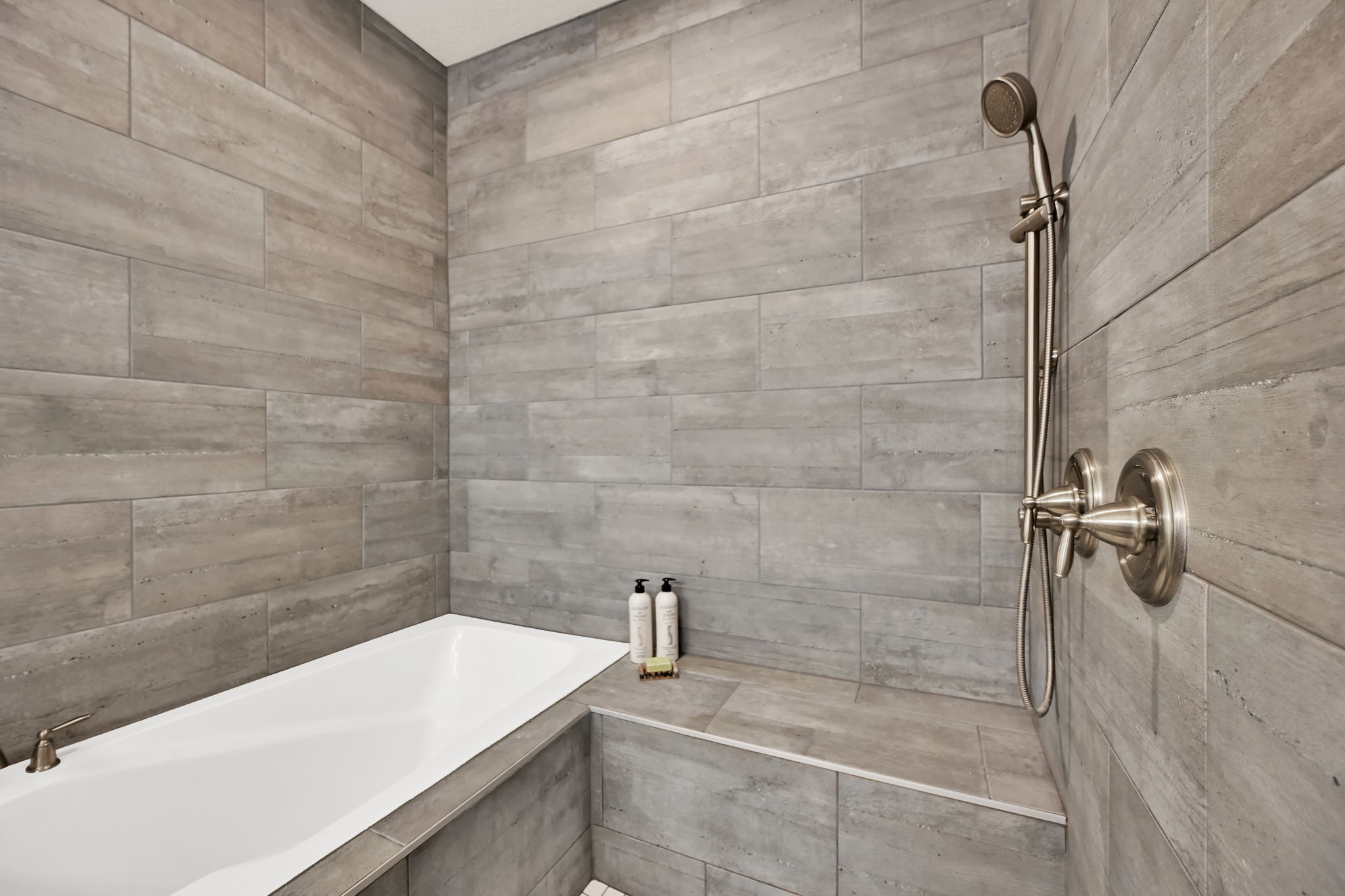 Bathtub and shower combination featuring floor-to-ceiling grey tile work, a built-in bench, and brushed nickel fixtures