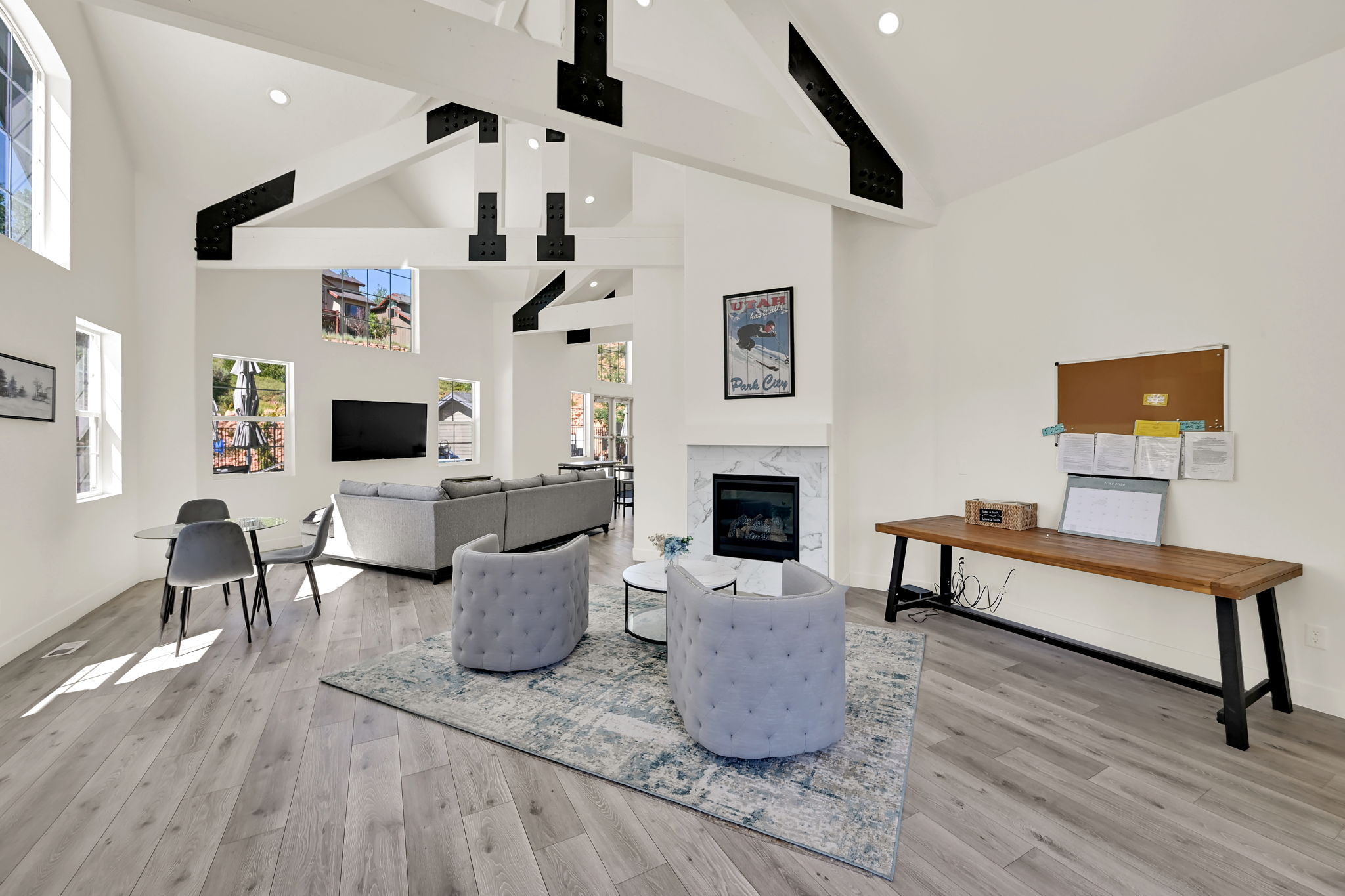 Expansive living area featuring a vaulted ceiling with exposed black beams, light wood-finish flooring, a contemporary fireplace with a marble-look surround, and multiple windows allowing ample natural light