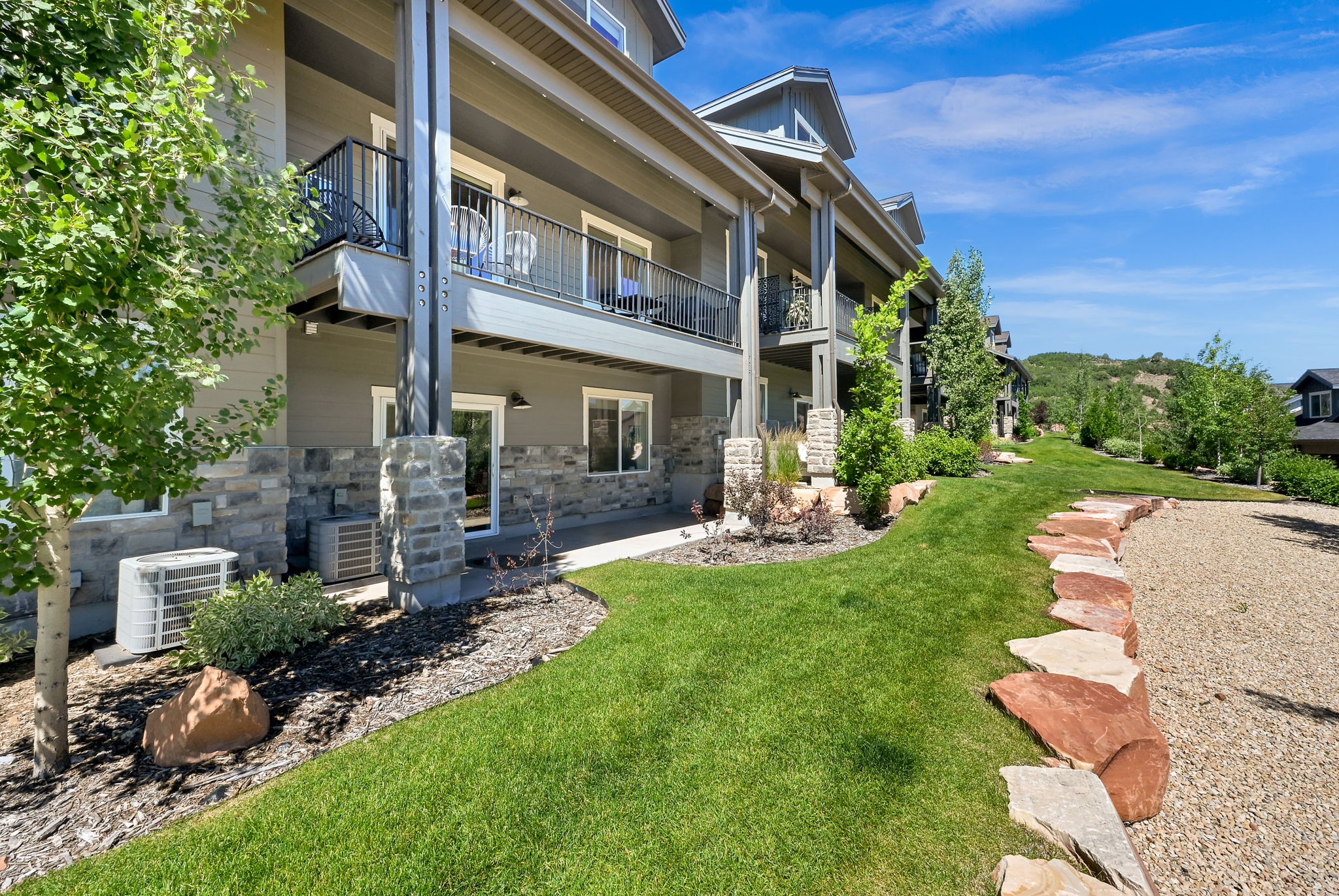 Multi-story building featuring stone veneer accents, individual balconies with iron railings, ground-level patios, and a landscaped lawn with rock borders