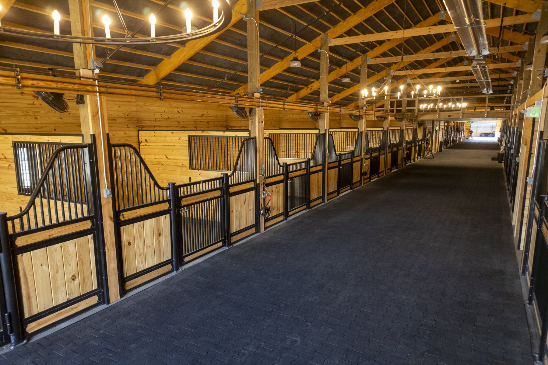 Barn interior featuring multiple stalls with wood and black metal construction, exposed wood beam ceiling, overhead lighting, and a dark-toned textured floor