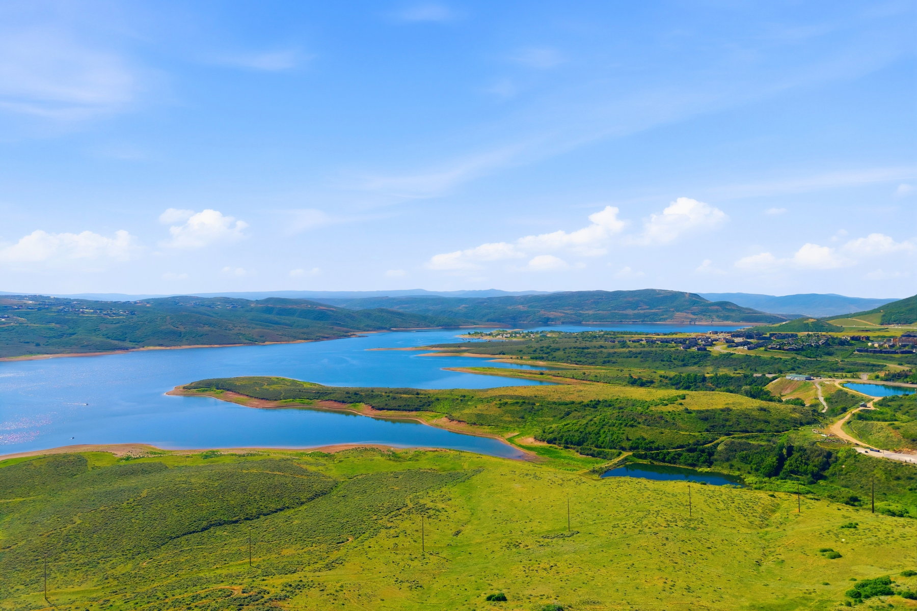 Expansive natural landscape featuring a large body of water, rolling green hills, and distant mountain ranges under a clear blue sky