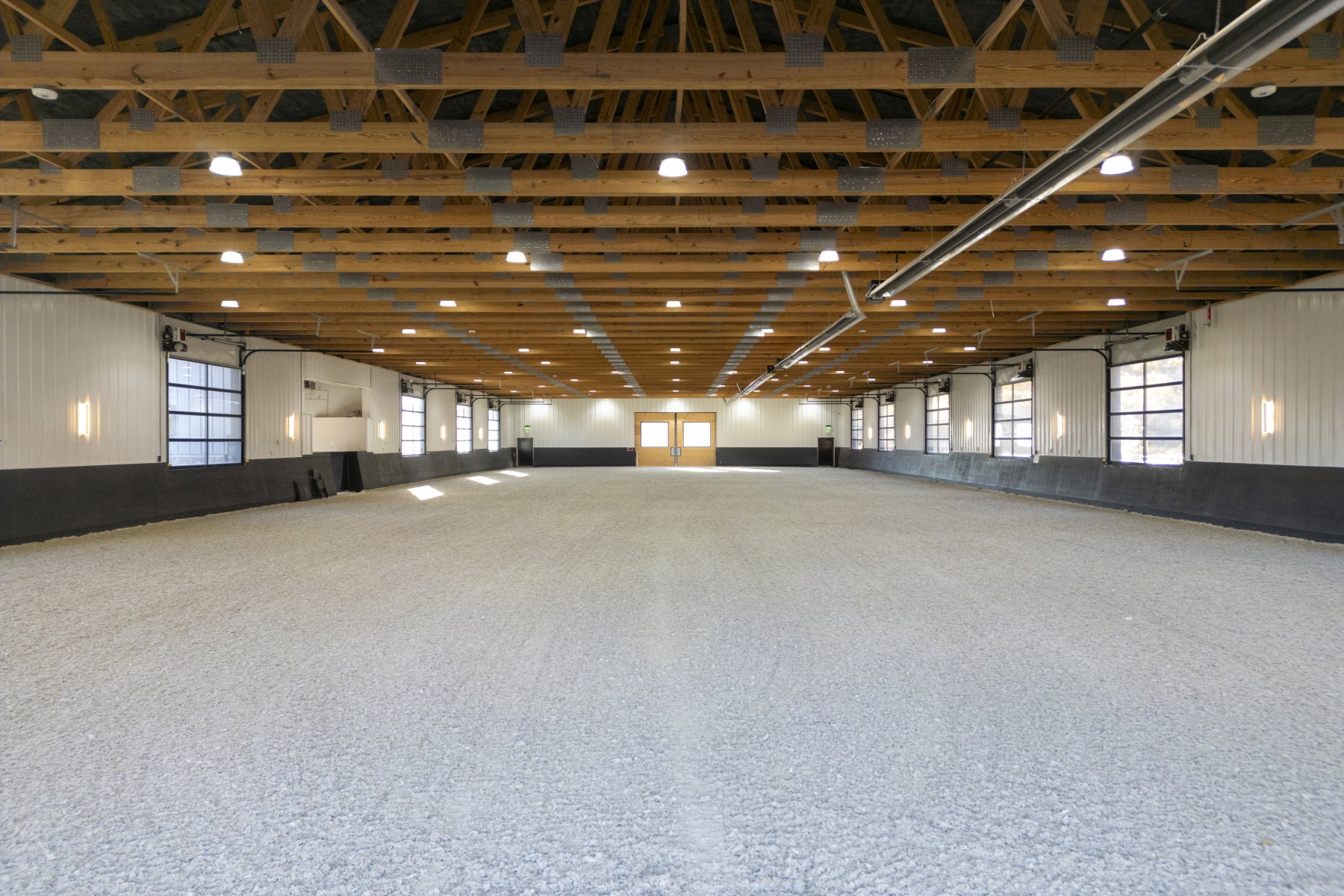 Spacious indoor arena featuring exposed wood beams, bright overhead lighting, and multiple roll-up garage doors with glass panels