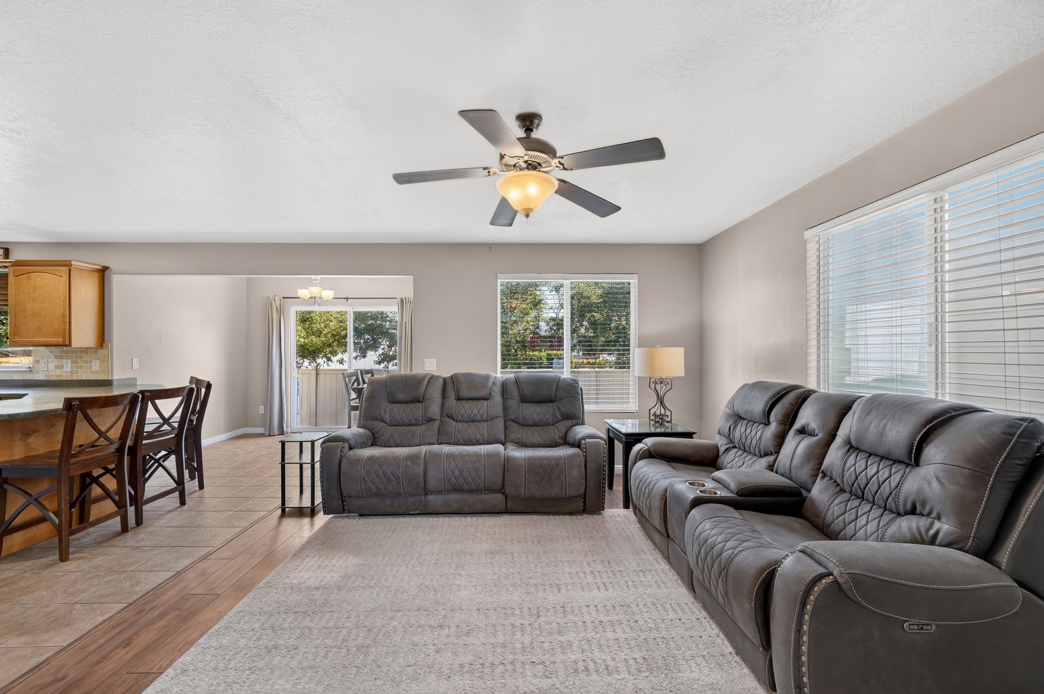 Expansive main living area featuring a ceiling fan with integrated lighting, multiple windows with blinds, a sliding glass door, and a combination of wood-finish flooring and tile