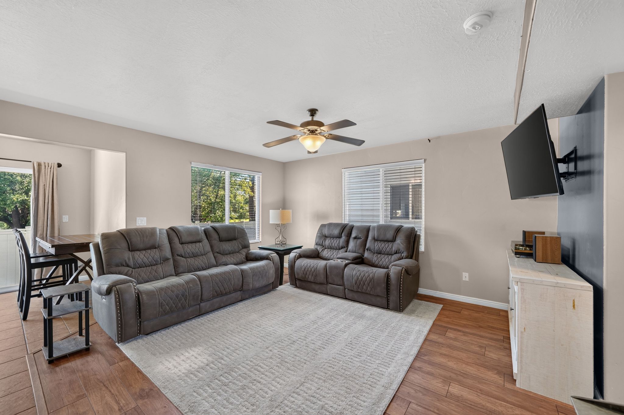 Open-concept living area featuring wood-finish flooring, a ceiling fan with integrated lighting, and multiple windows