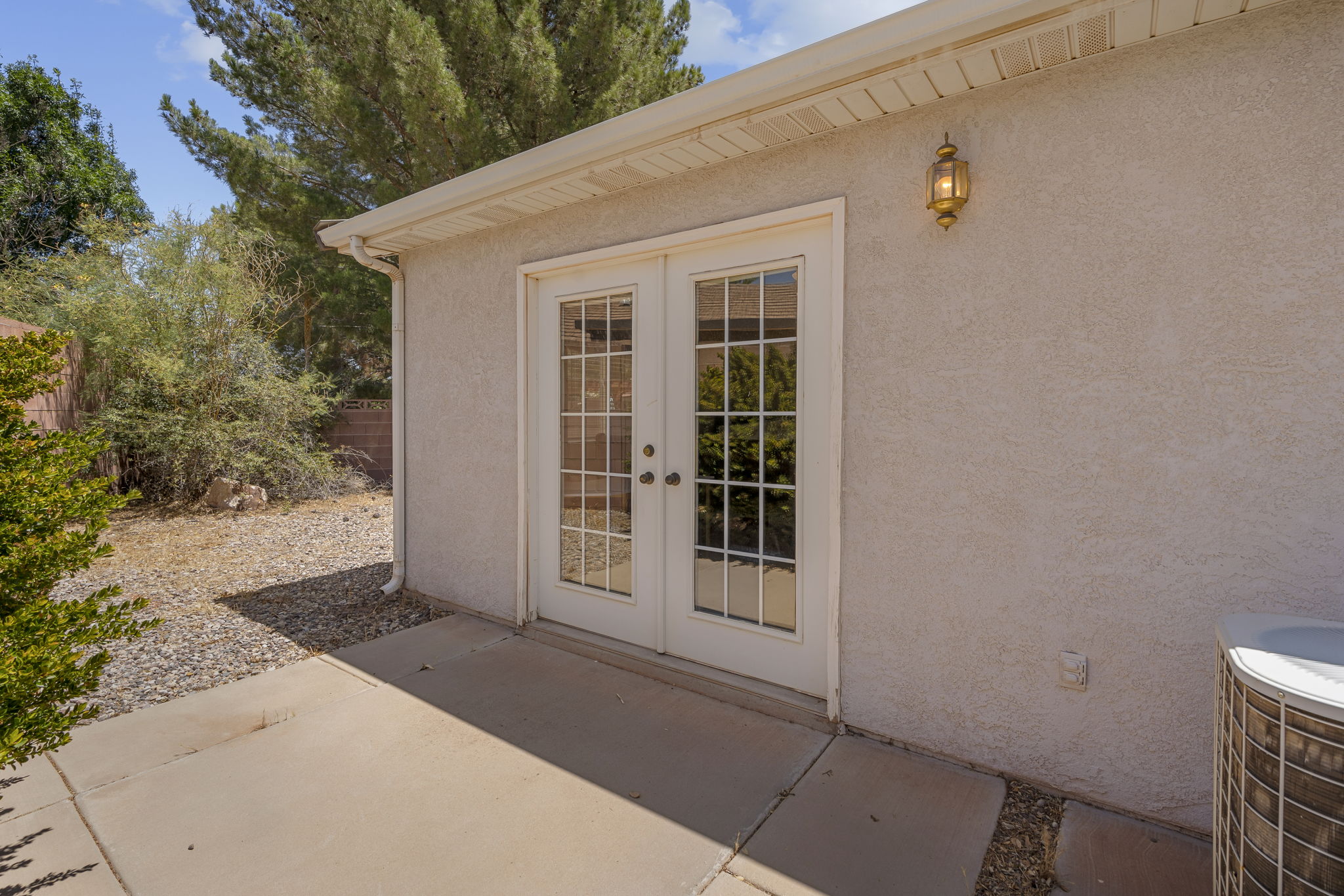 Exterior patio area featuring a textured stucco facade, white French doors with divided lites, a wall-mounted brass lantern fixture, and a concrete patio pad