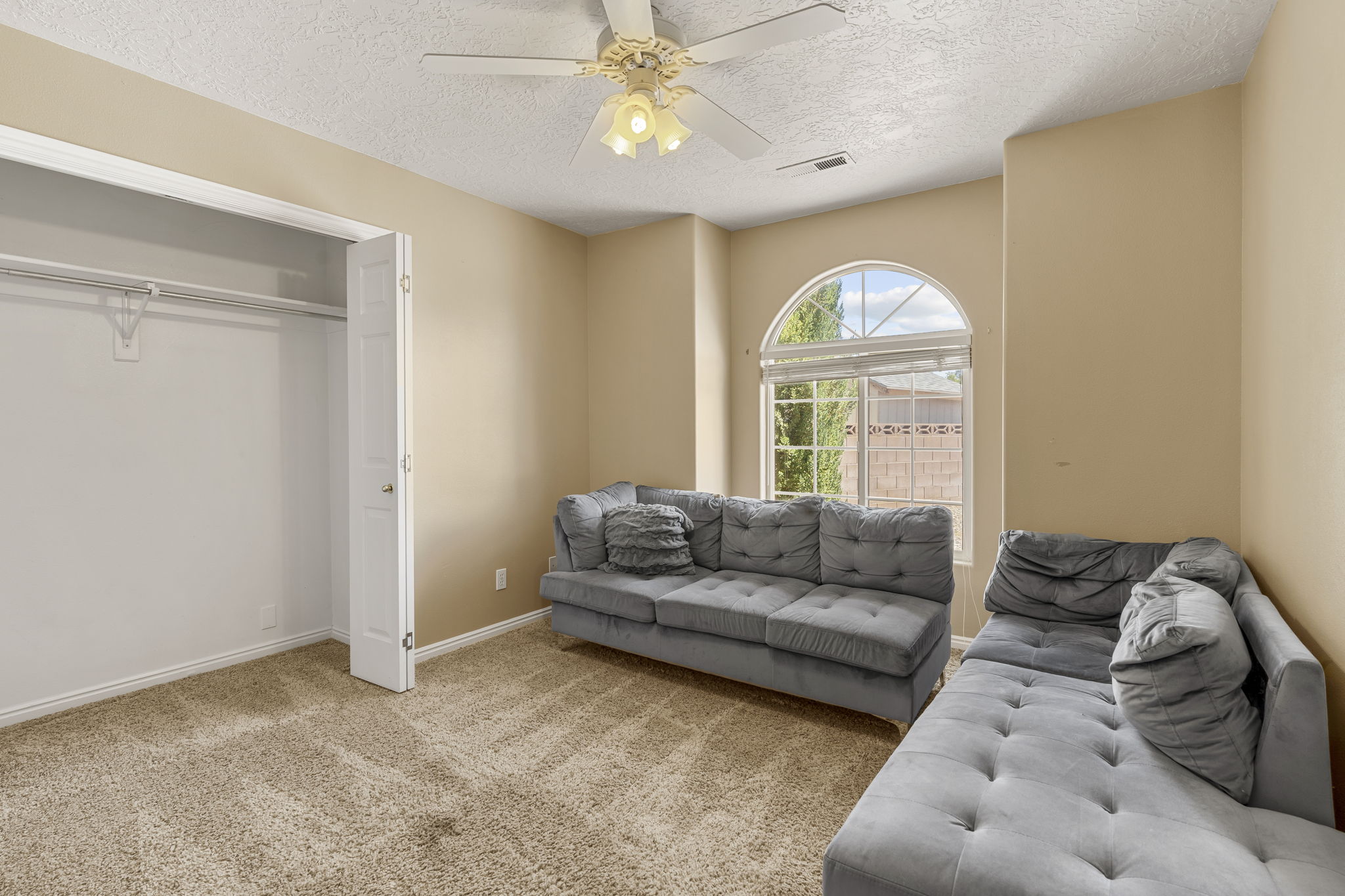 Room featuring a large arched window with horizontal blinds, textured beige carpeting, a built-in closet with bifold doors and hanging rod, a ceiling fan with light fixture, and neutral-toned walls