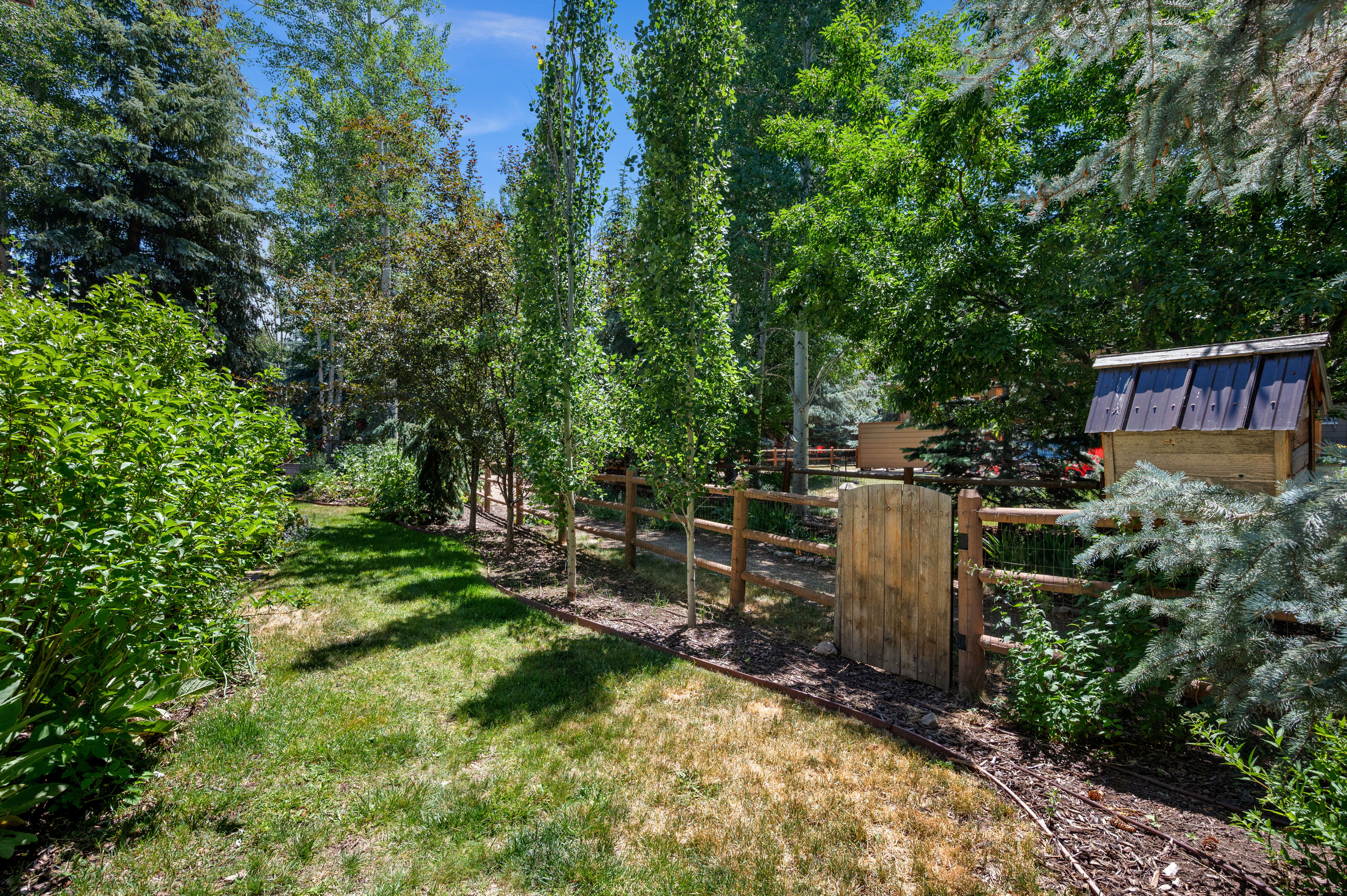 Fenced backyard featuring a wooden gate, split-rail fencing, mature trees, and established landscaping