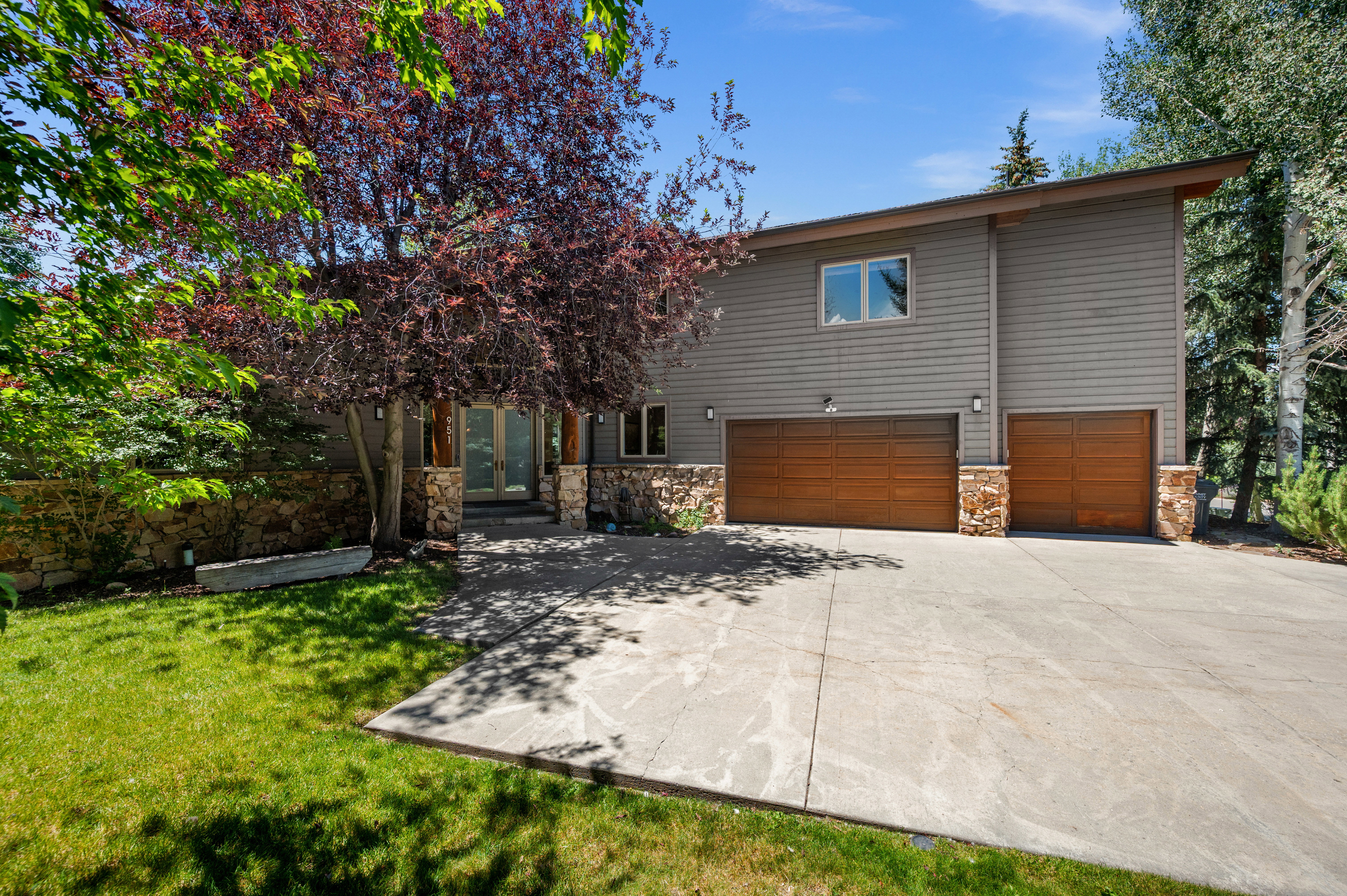 Contemporary exterior with gray horizontal siding, natural stone accents, two wood-finish garage doors, a concrete driveway, and mature landscaping