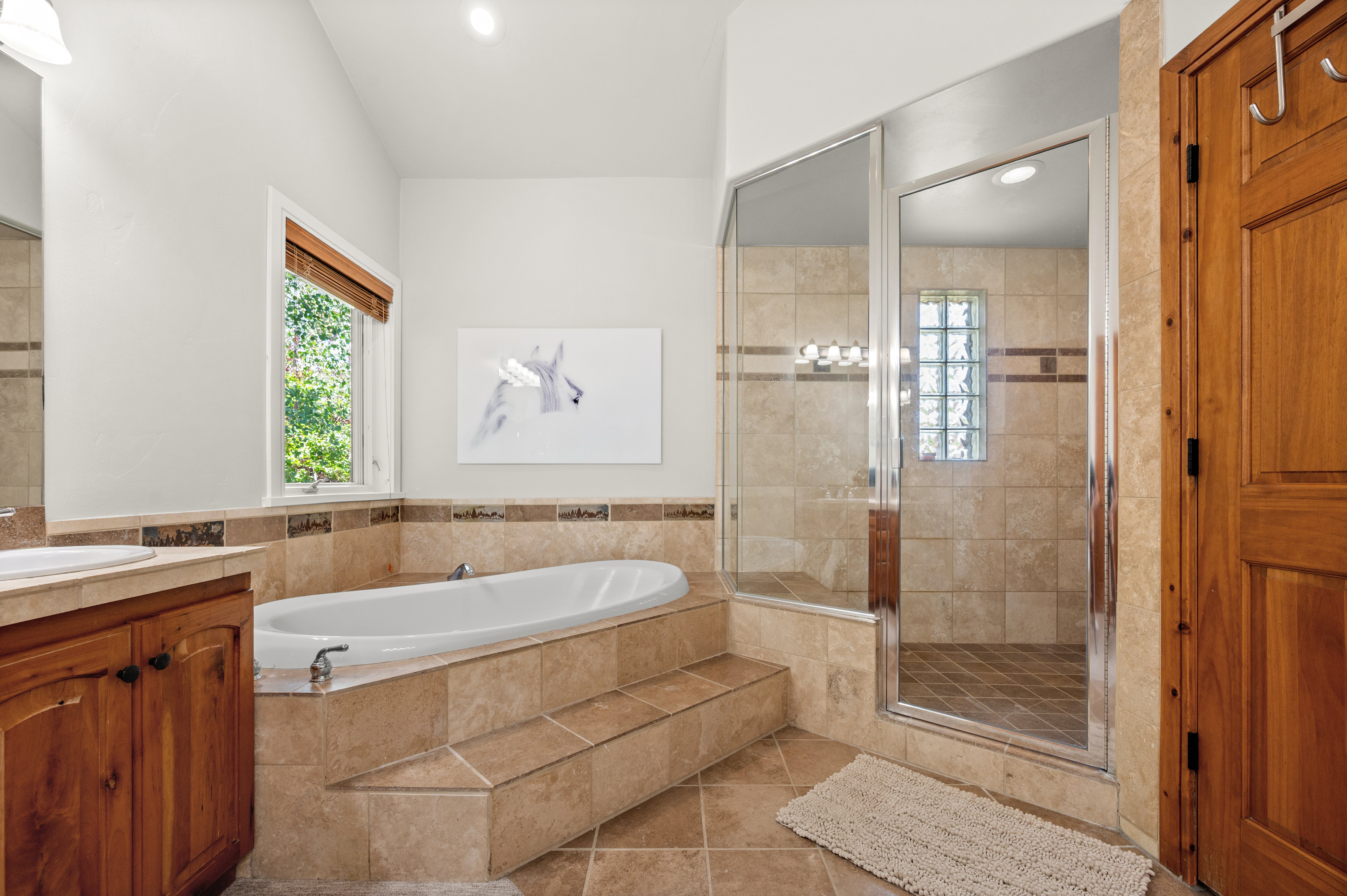 Spacious bathroom featuring a built-in soaking tub with tiled surround, a separate glass-enclosed shower with tiled walls and a glass block window, and dual vanities with wood cabinetry and undermount sinks