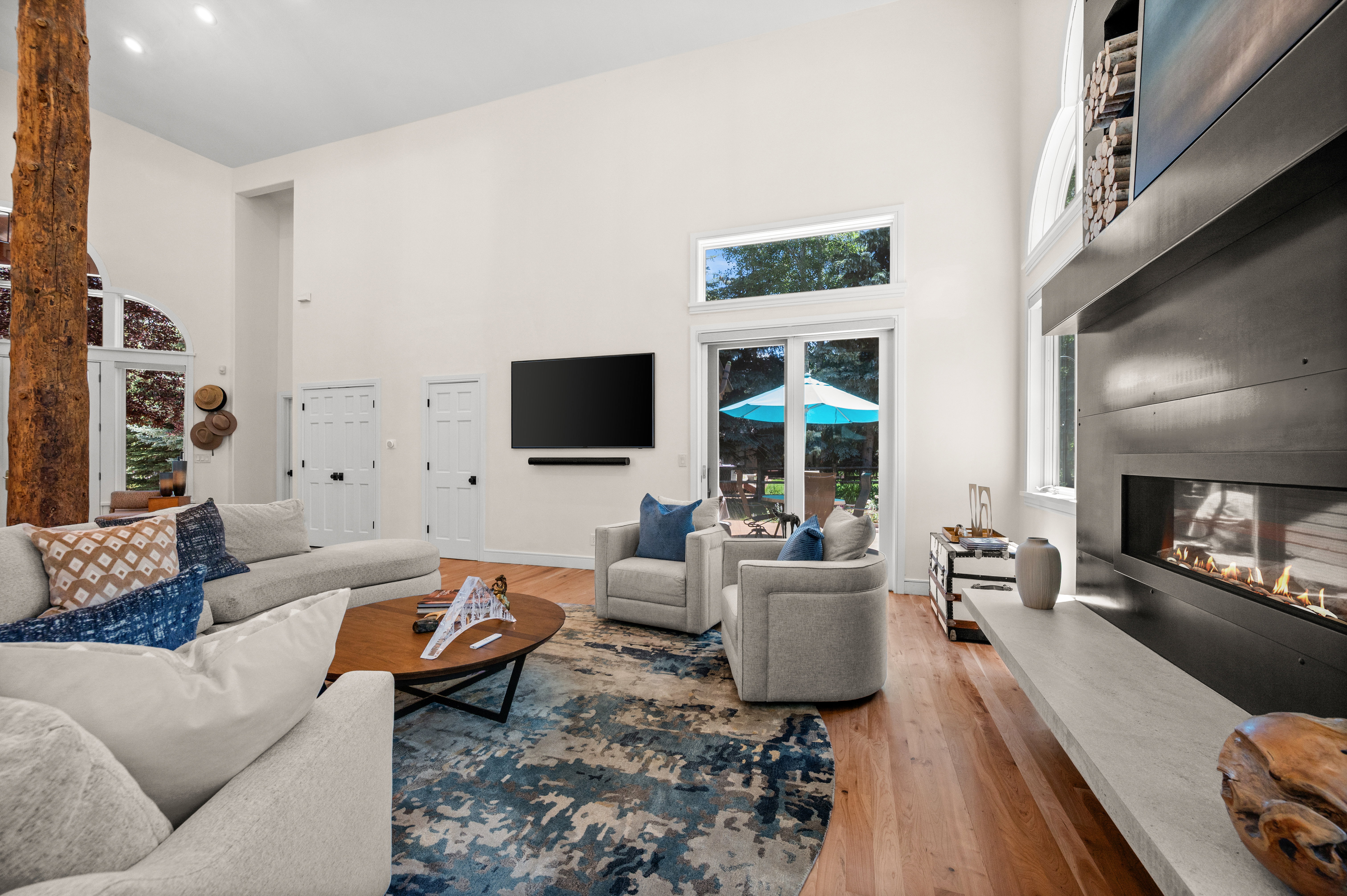 Spacious living area featuring high ceilings, hardwood flooring, a prominent tree-trunk architectural feature, a contemporary fireplace with a wide hearth, and glass French doors leading to exterior grounds