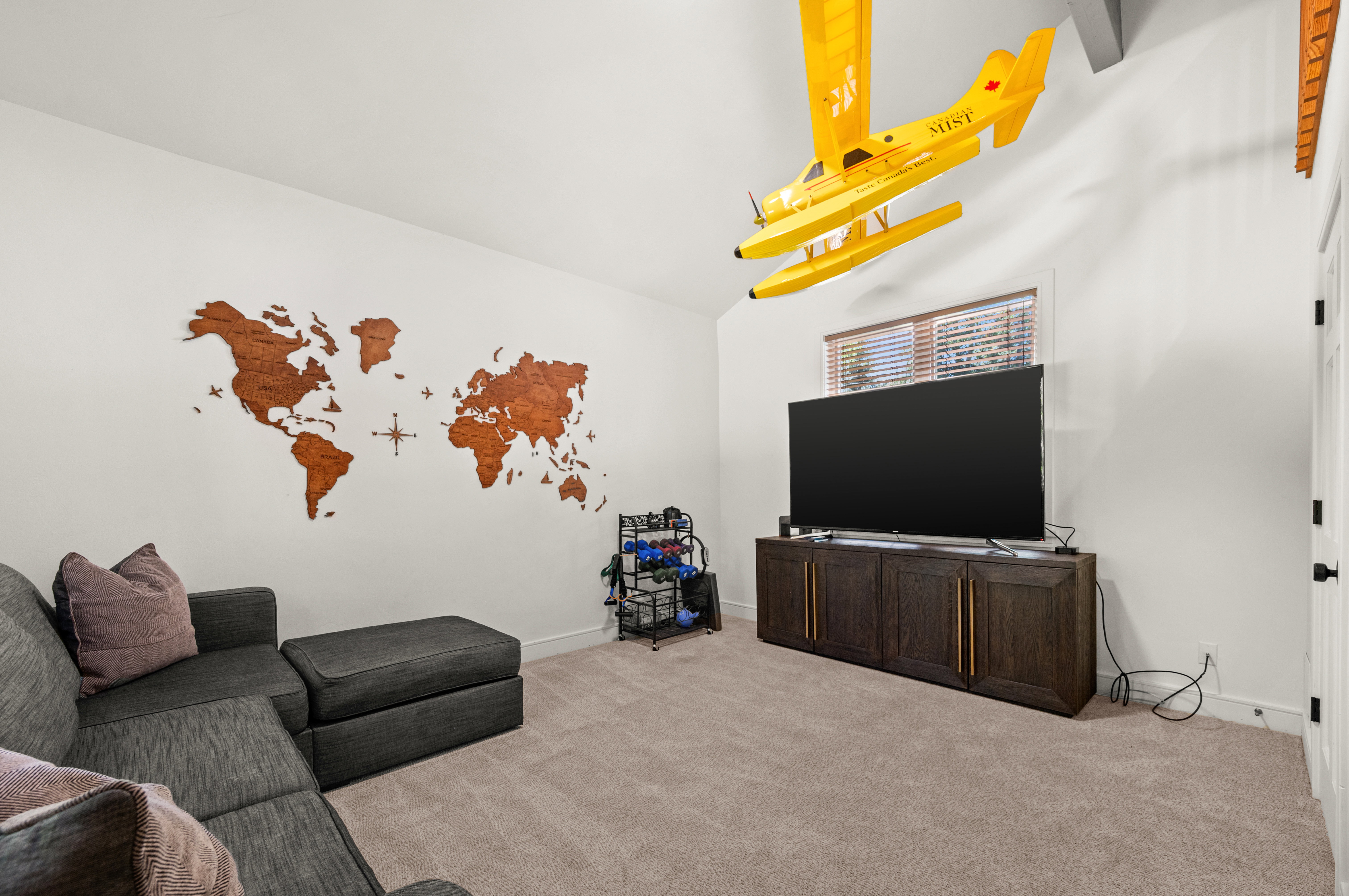 Carpeted room featuring a white wall with a wooden world map art piece, a window with horizontal blinds, and a built-in media console with cabinet storage