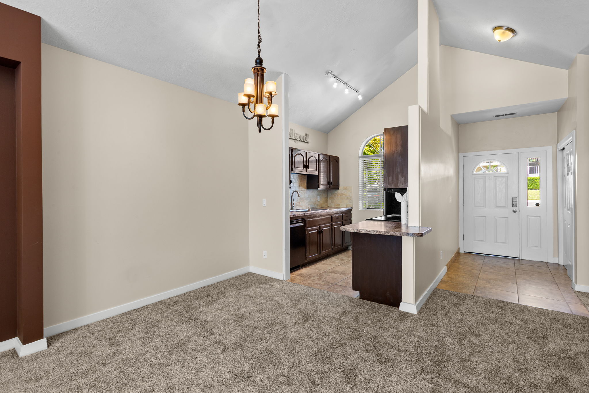 Vaulted ceilings highlight this open-concept interior, featuring a chandelier, carpeted living area, and a kitchen with dark cabinetry and a breakfast bar