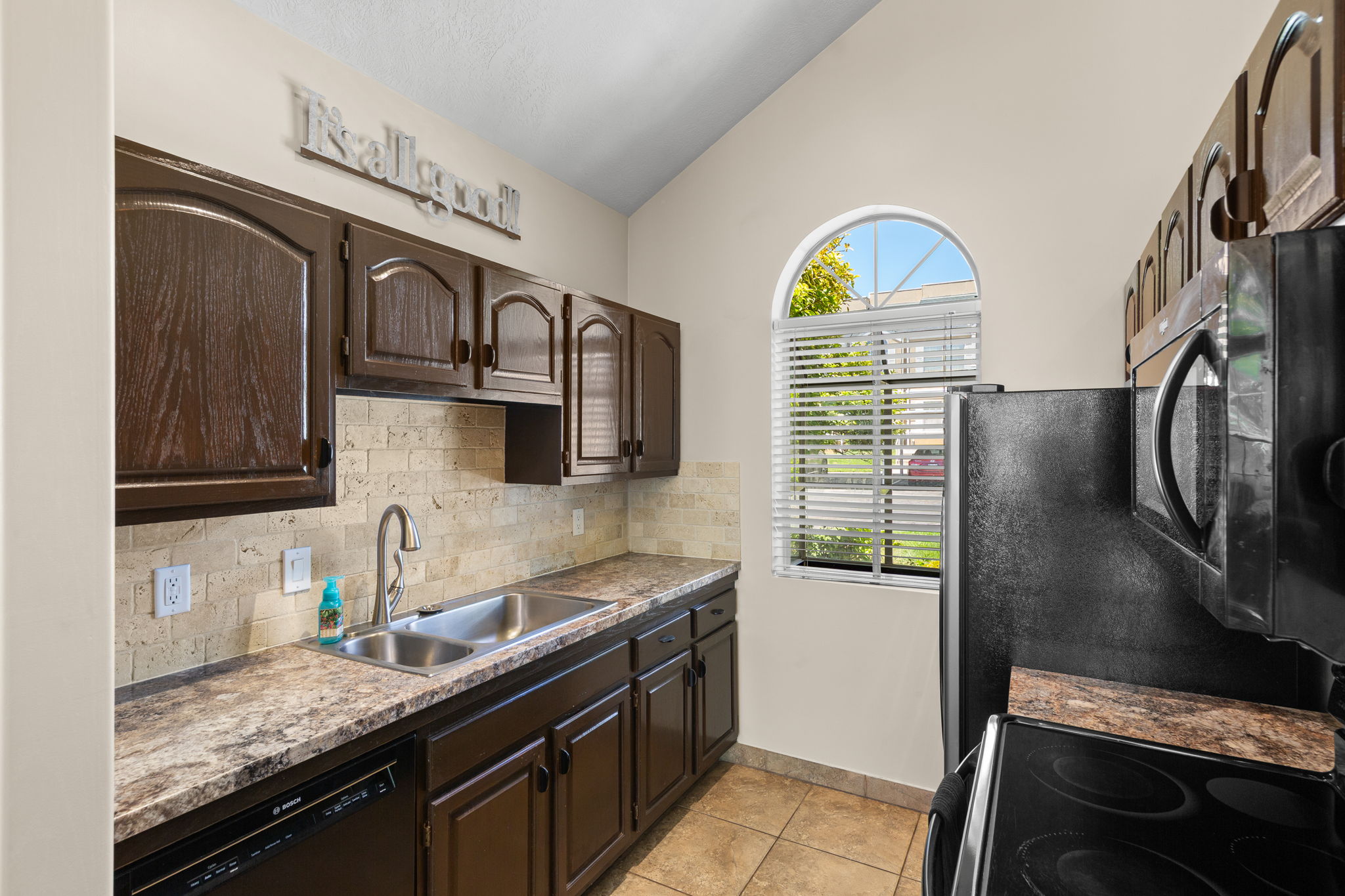 Kitchen featuring dark wood cabinetry, a double basin stainless steel sink, and a light-toned stone tile backsplash