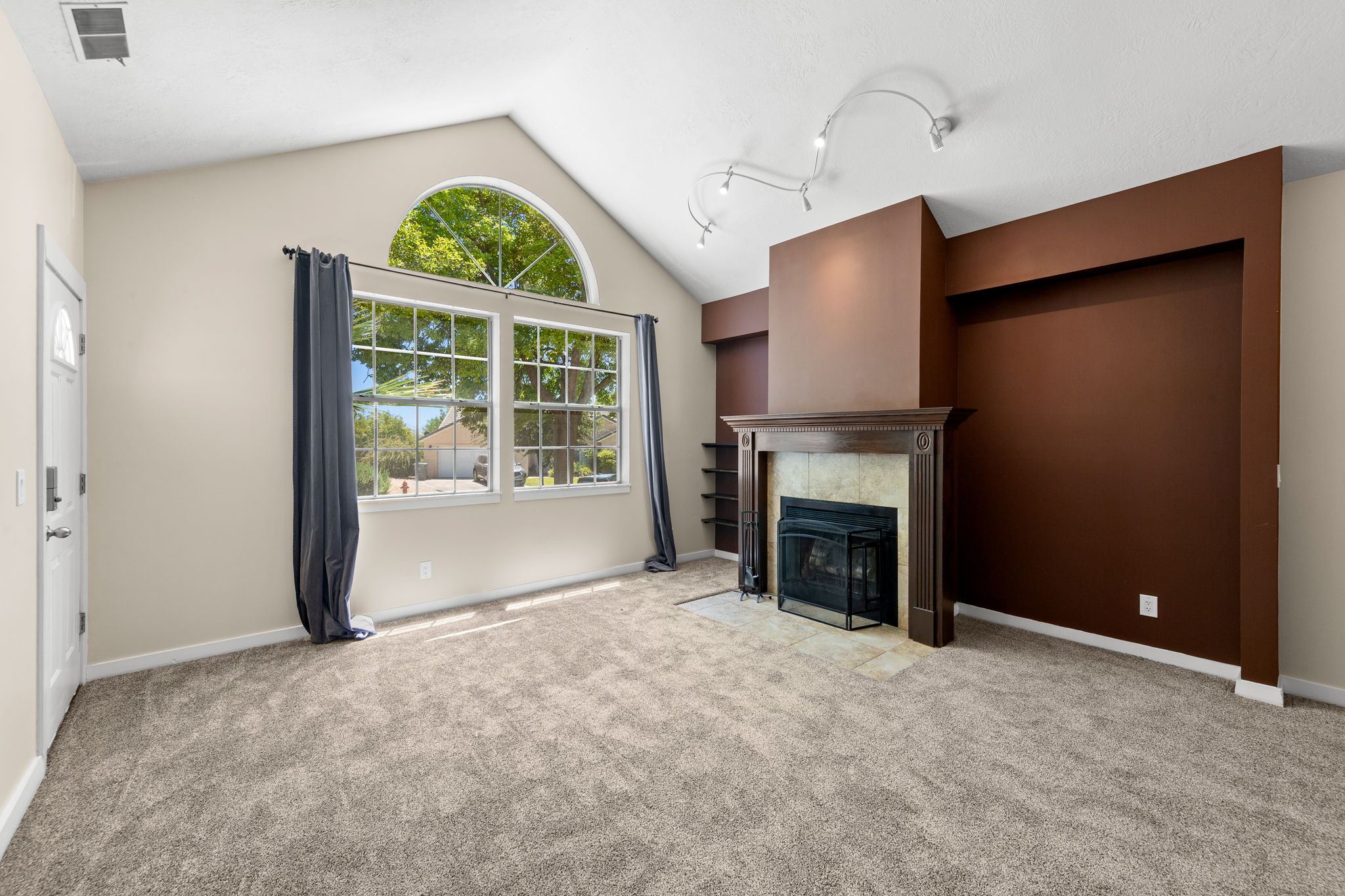 Living area featuring vaulted ceilings, a prominent fireplace with wood-finish mantel and tile surround, large windows with an arched transom, track lighting, and soft gray carpeting