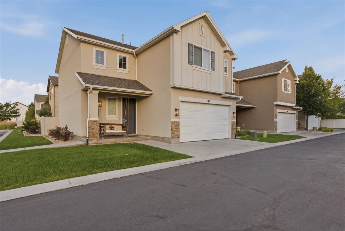 Contemporary stucco exterior with stone accents, a covered entry porch, an attached two-car garage, manicured lawn, and mature landscaping