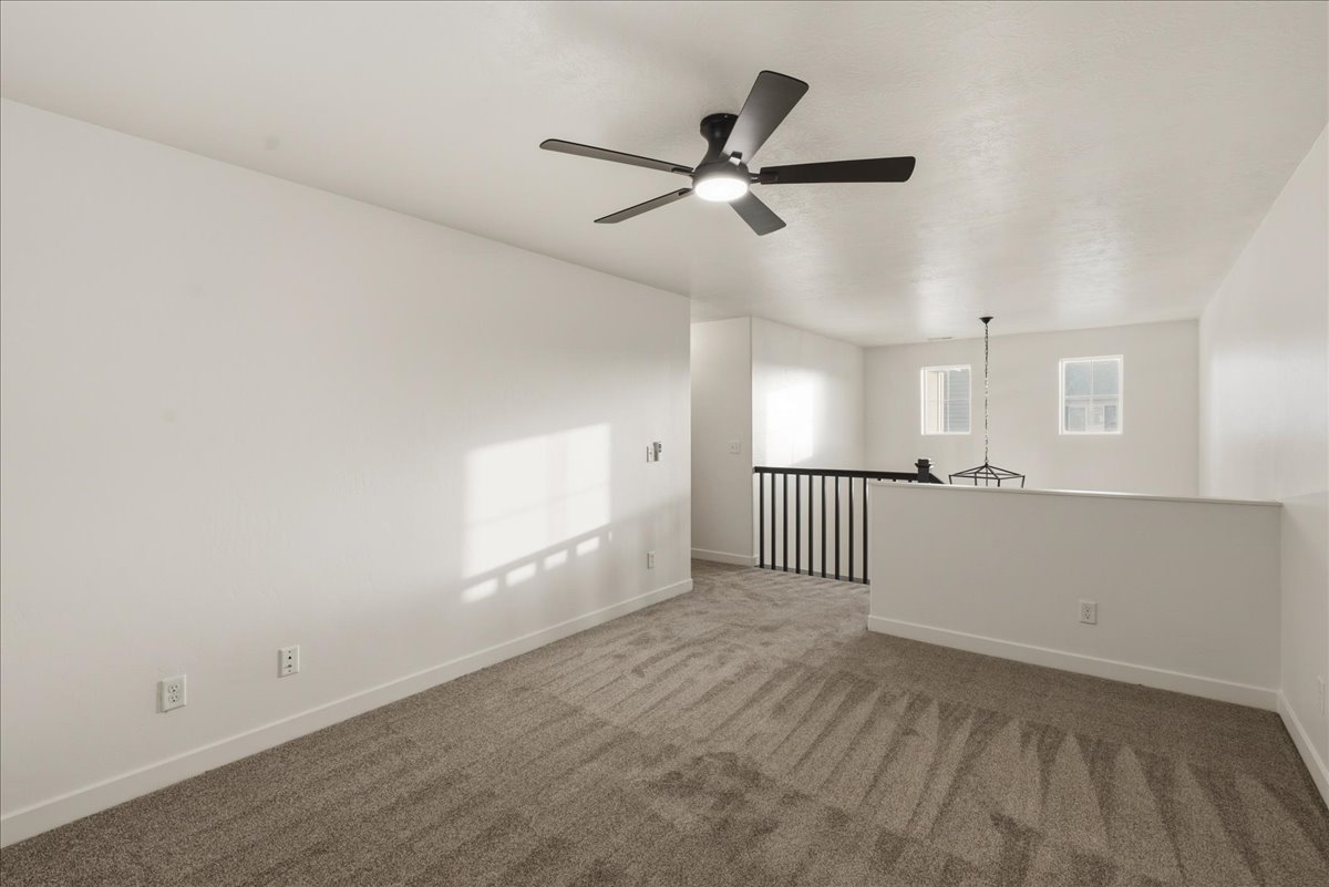 Expansive upper-level space featuring a contemporary ceiling fan, plush carpet flooring, white walls, and a black metal railing overlooking the lower level