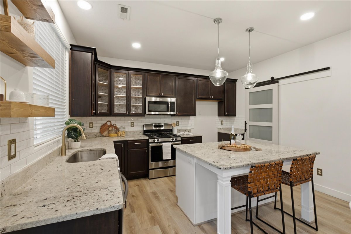 Open-concept kitchen featuring dark wood-finish cabinetry, stainless steel appliances, light-toned countertops, and a central island with pendant lighting