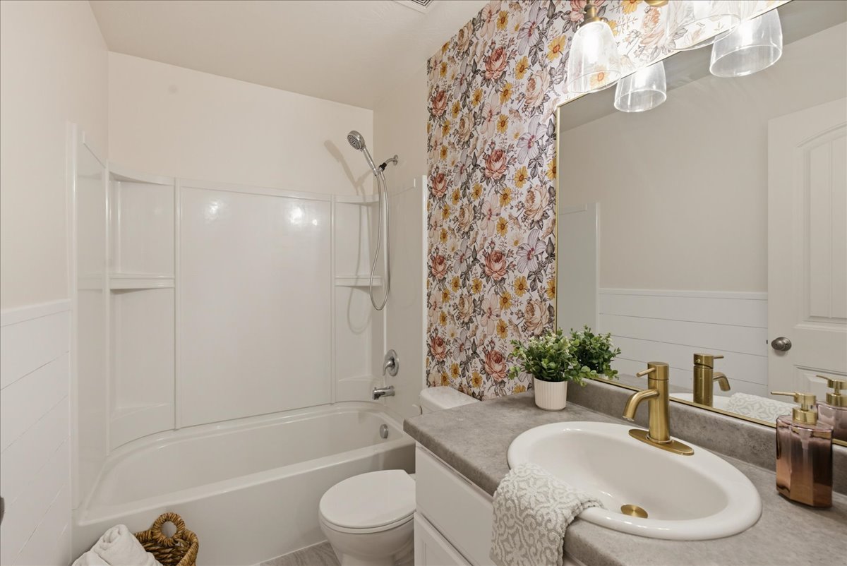 Bathroom featuring a white tub and shower combination, a single vanity with an integrated sink, a gold-tone faucet, a mirror, and floral accent wallpaper