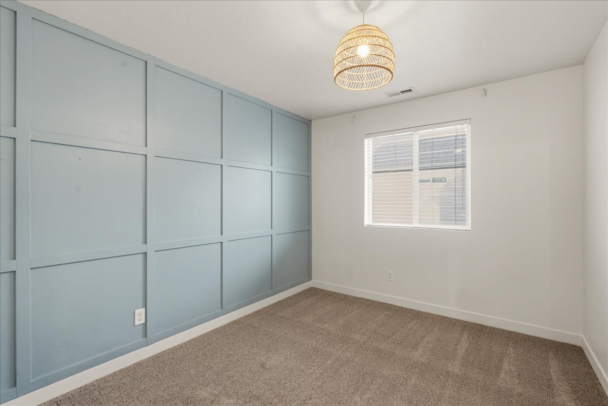 Bedroom featuring a light blue accent wall with decorative paneling, neutral carpeting, a single window with blinds, a white ceiling, and a woven pendant light fixture