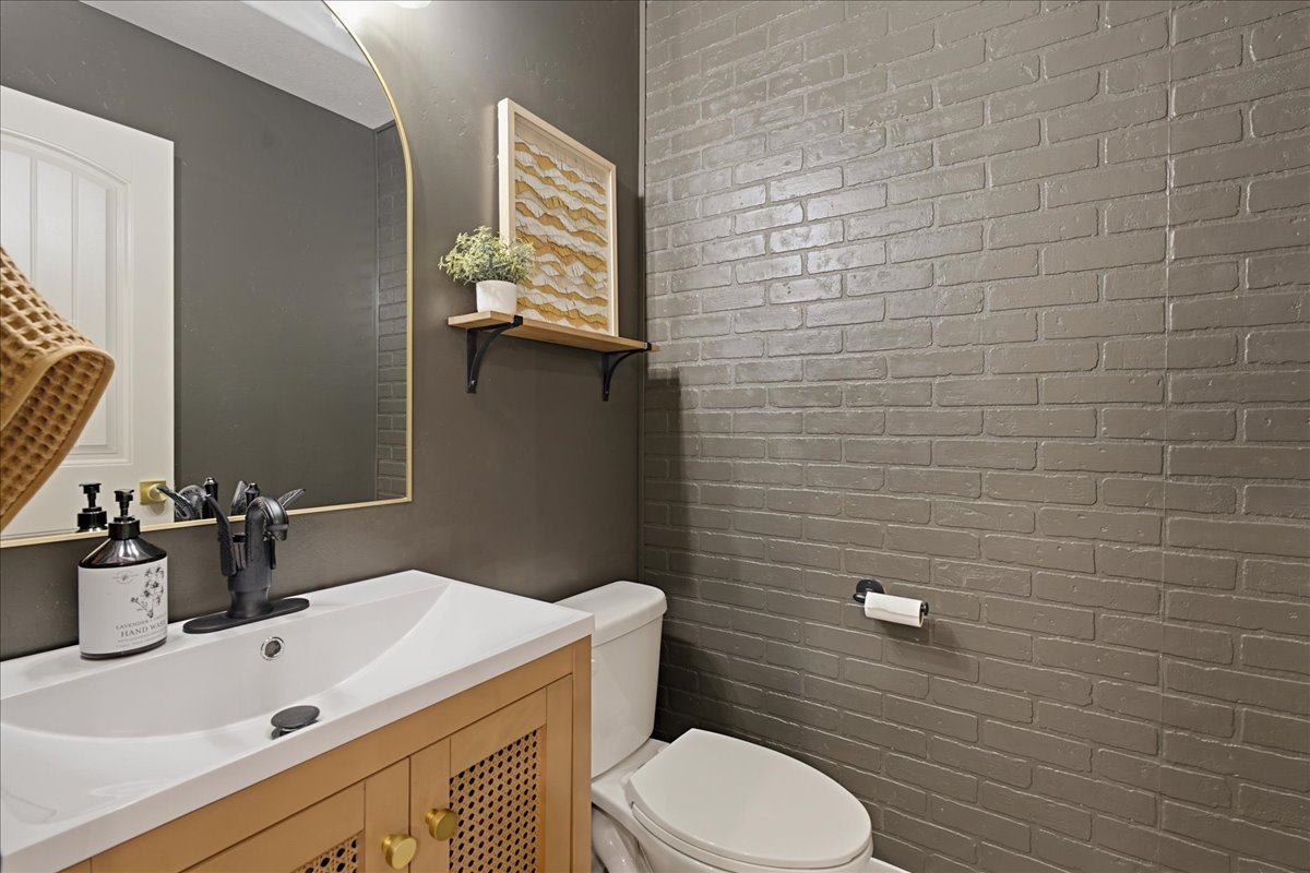 Bathroom featuring a light wood-finish vanity with woven cabinet fronts, a white integrated sink, a rectangular gold-frame mirror, and a painted brick accent wall