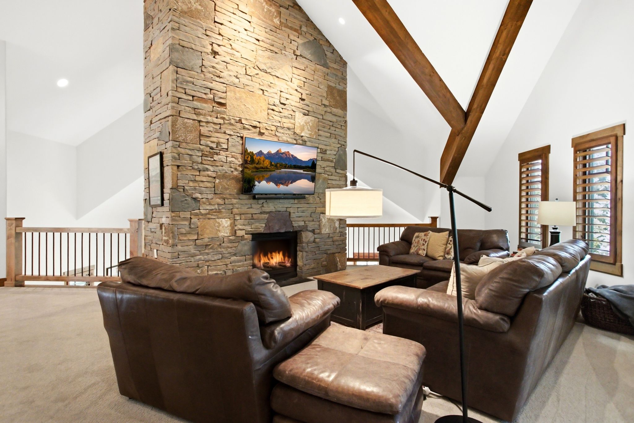 Vaulted ceiling living area featuring a floor-to-ceiling stone fireplace, exposed wood beams, plantation shutters, and light wood railing with black spindles