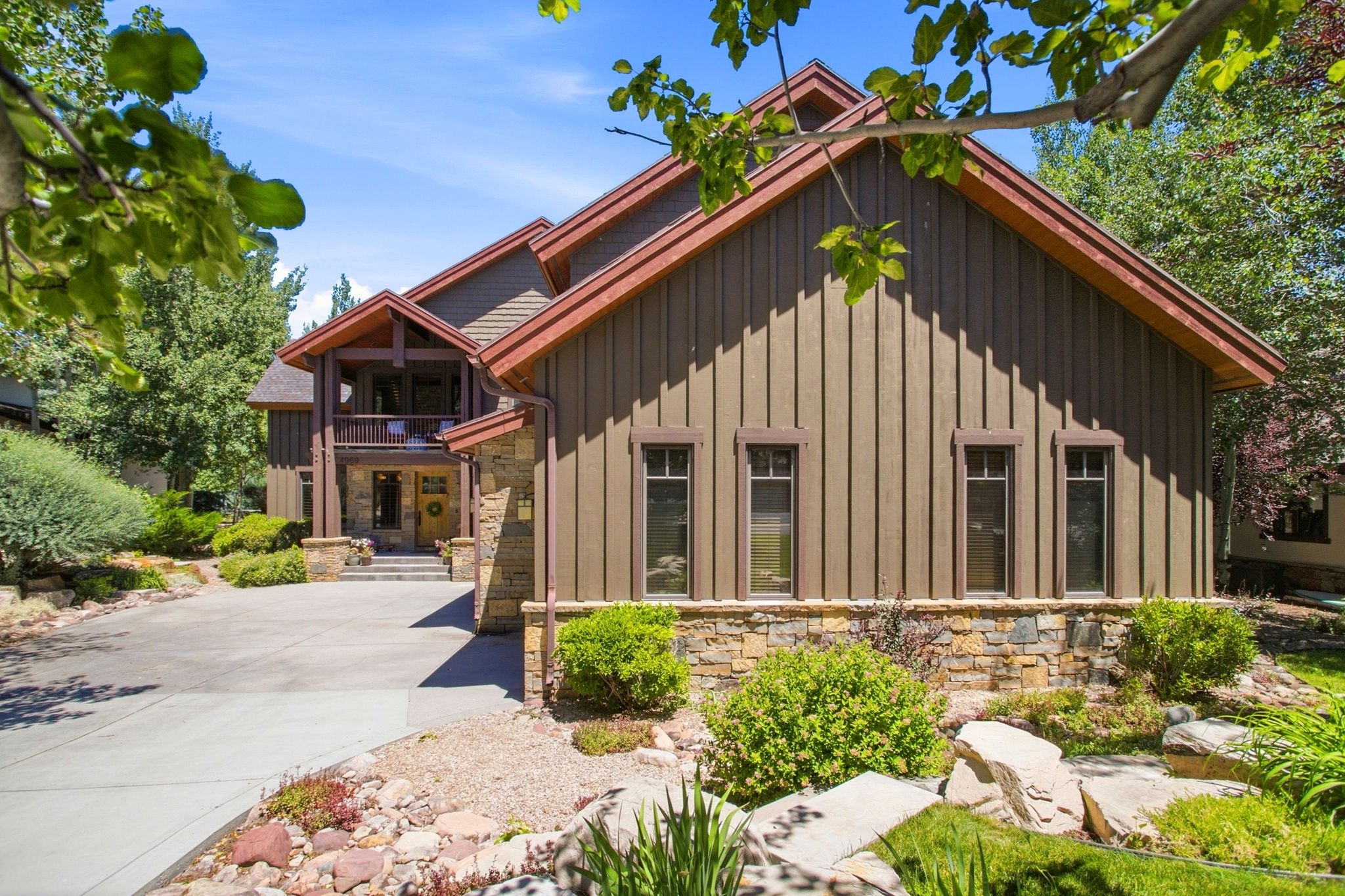 Stone and board-and-batten exterior featuring a prominent gabled roofline, a covered entrance with an upper-level balcony, and a paved driveway leading to the main entrance