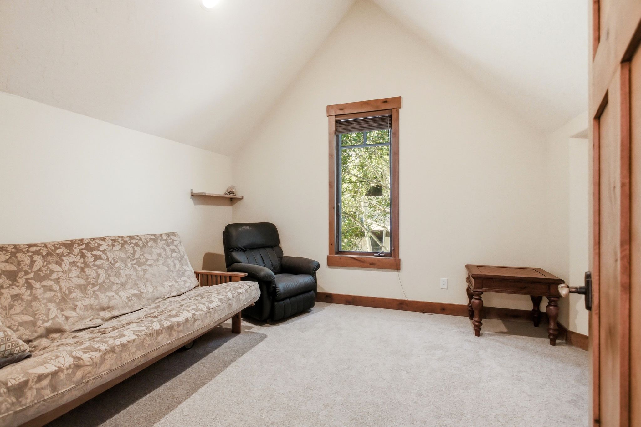 Vaulted ceiling room featuring a single window with wood trim, carpet flooring, a natural wood paneled door, and a wall-mounted shelf