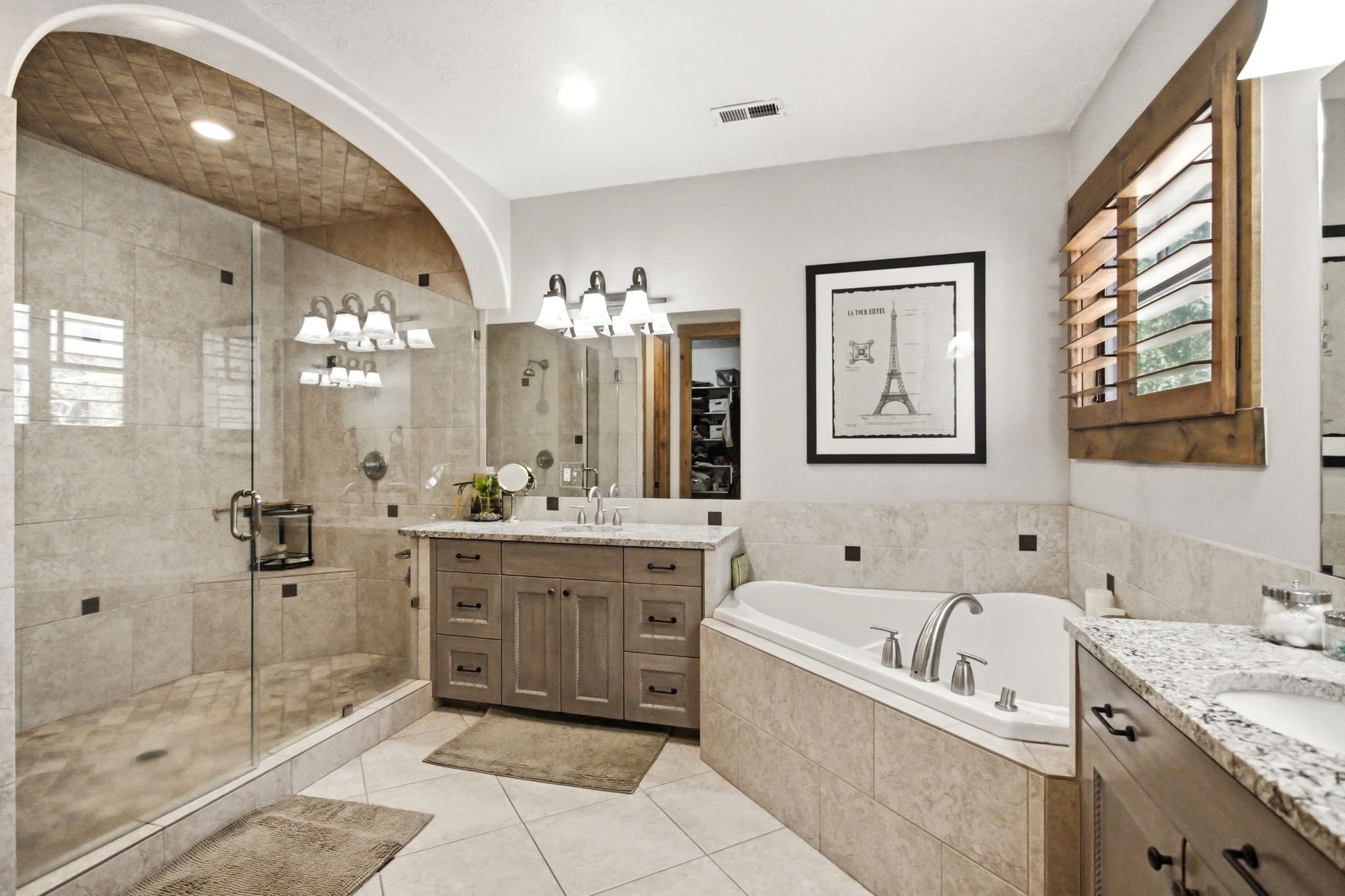 Spacious bathroom featuring a glass-enclosed shower with a tiled archway and textured ceiling, a corner soaking tub, and dual vanity areas with granite-finish countertops and wood cabinetry