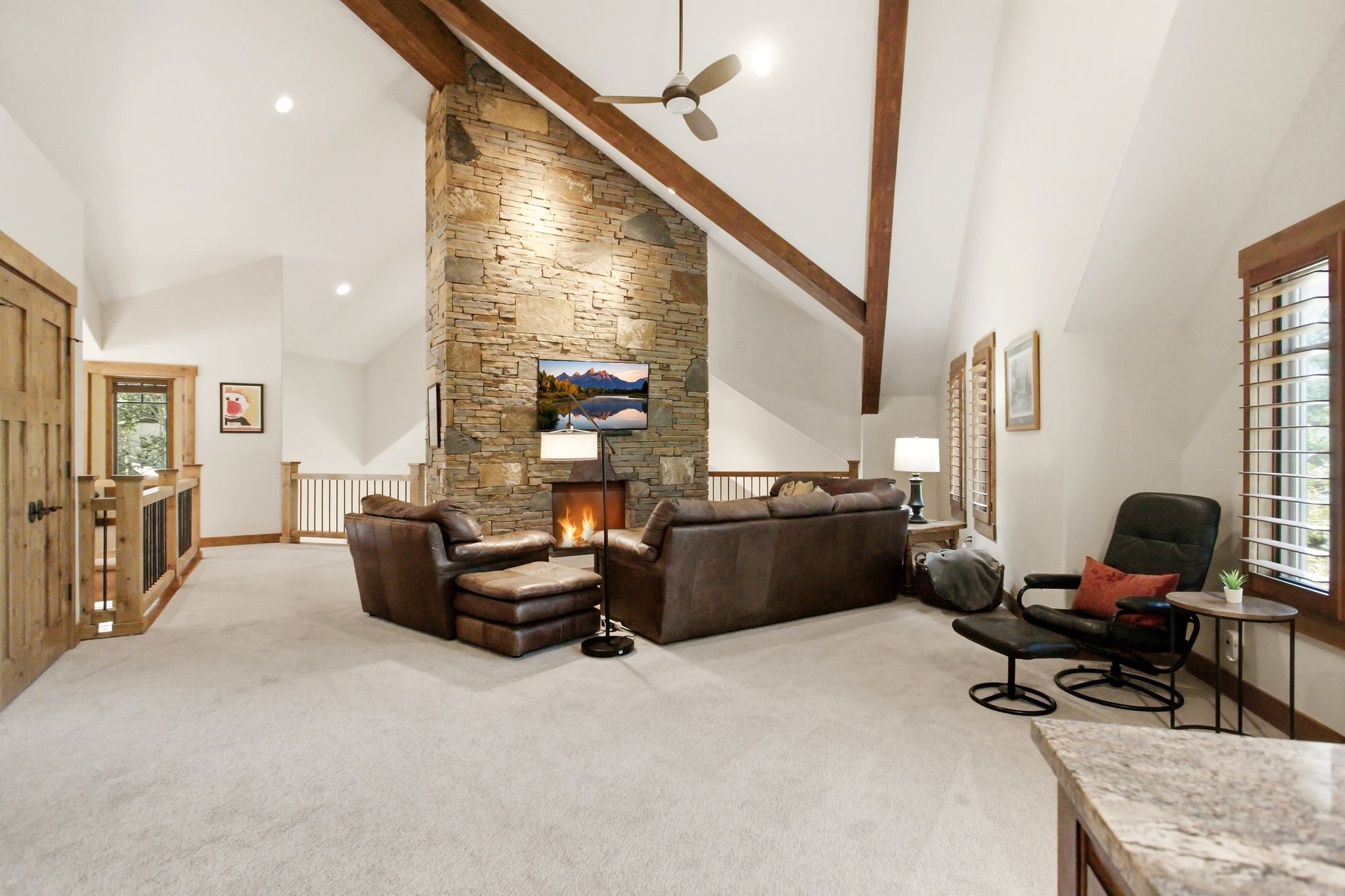 Spacious living area featuring a vaulted ceiling with exposed wood beams, a stone-clad fireplace, and neutral carpeting
