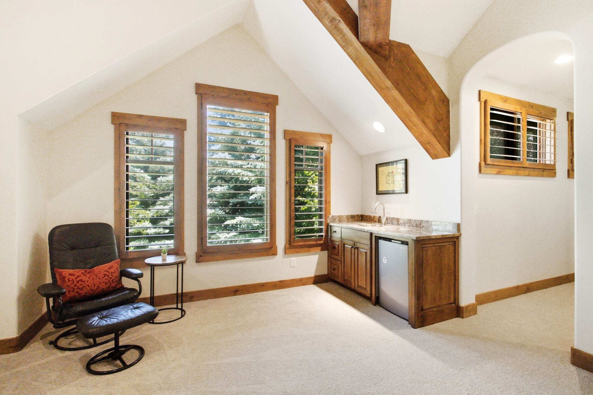 Accentuated by exposed wood beams, this space features multiple windows with wood-framed plantation shutters, a built-in wet bar with a countertop, under-cabinet storage, and a compact refrigerator