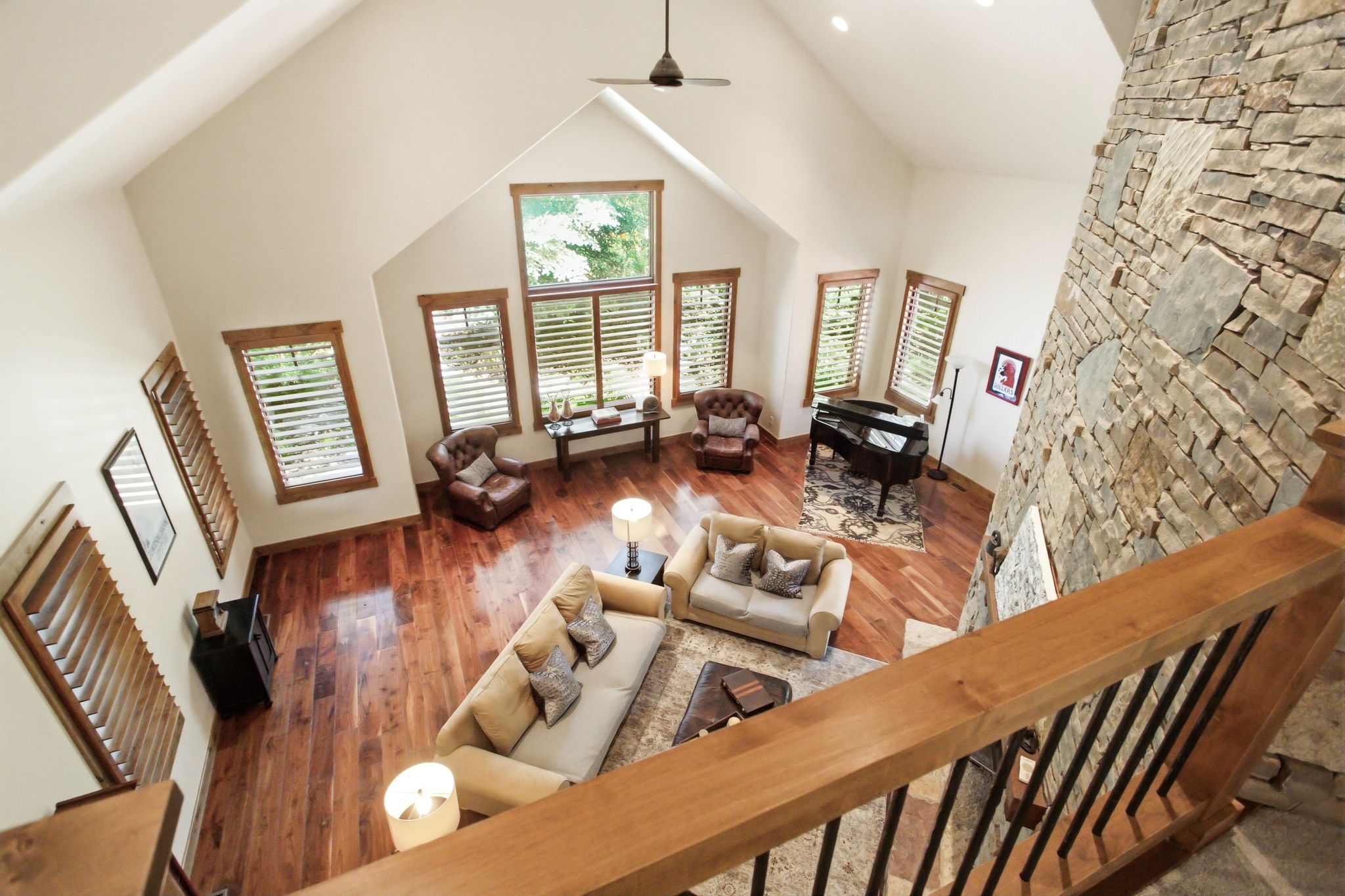 Vaulted ceiling great room featuring a floor-to-ceiling stone-clad fireplace, wood-finish flooring, multiple windows with plantation shutters, and an open-concept design