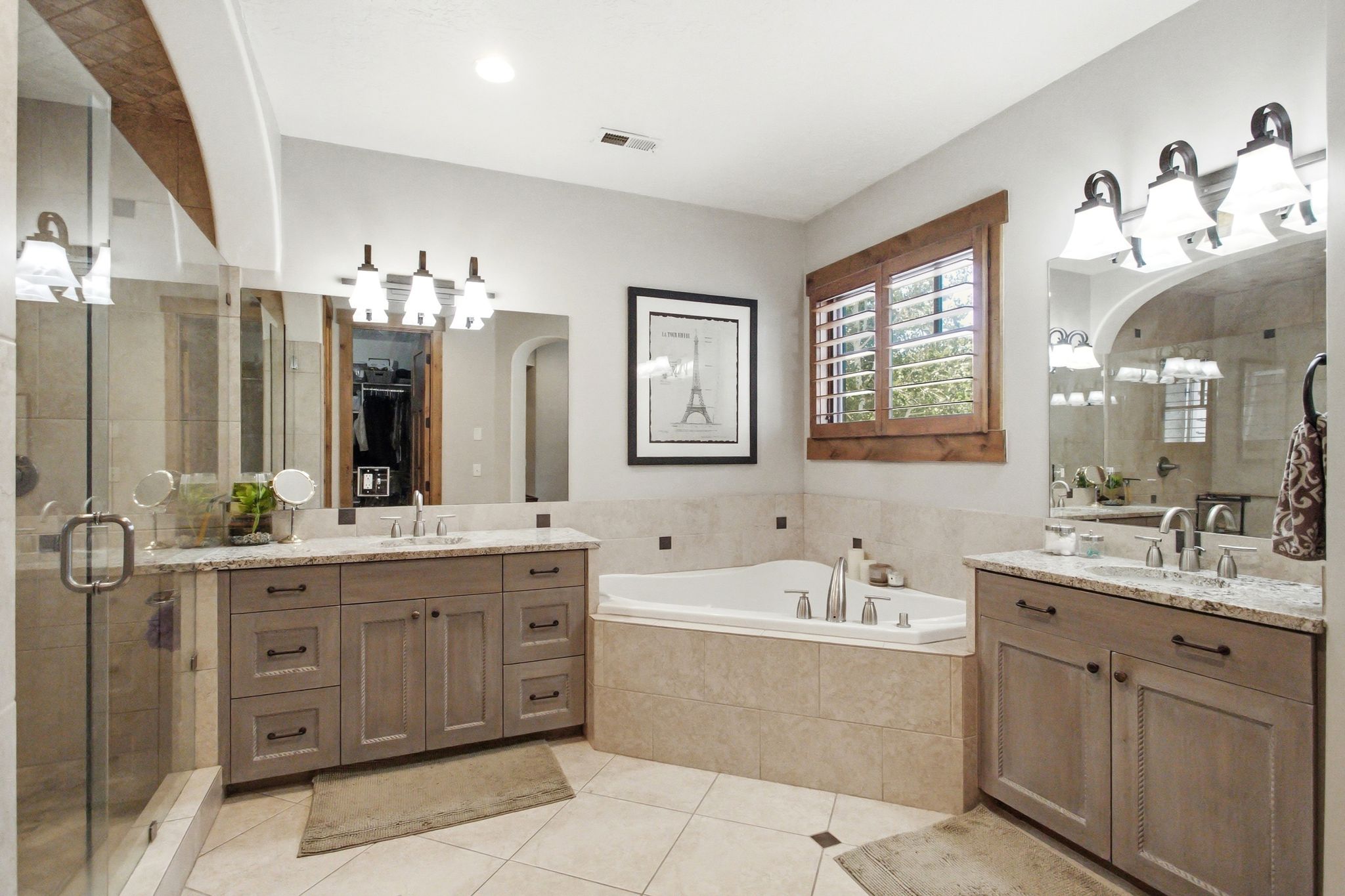 Spacious bathroom featuring a built-in corner soaking tub, a large glass-enclosed shower, dual vanities with granite countertops, and light wood-finish cabinetry