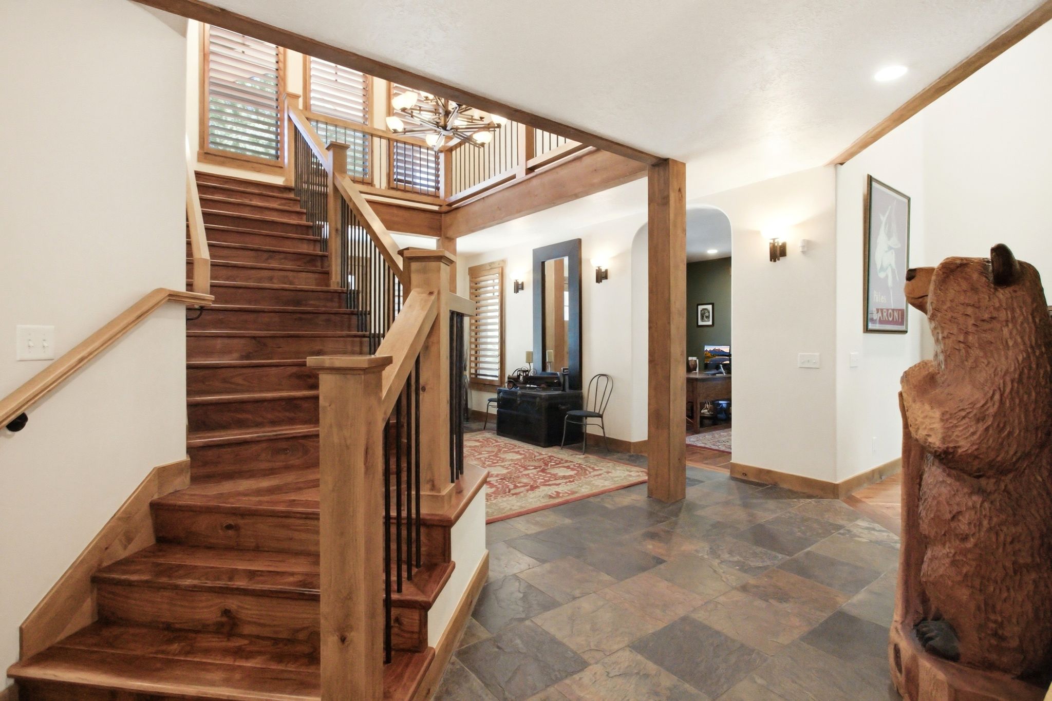 Grand entryway featuring wood-finish stairs with black metal spindles, slate tile flooring, exposed wood beams, and a dual-story ceiling with a chandelier