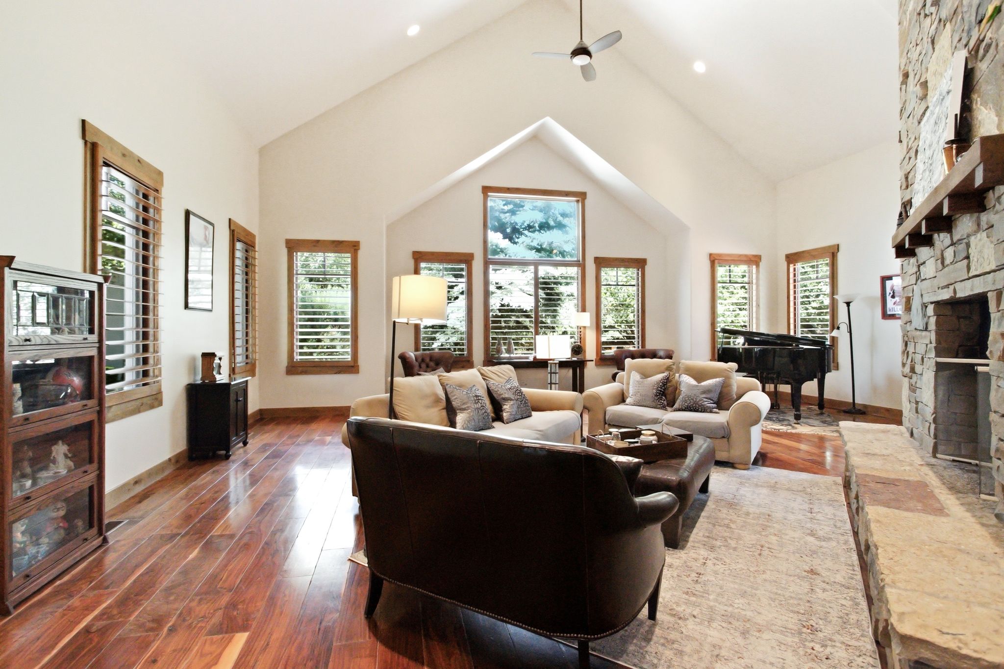 Vaulted ceiling with a ceiling fan and recessed lighting, wood-finish flooring, stone fireplace, multiple windows with white plantation shutters, and natural wood trim throughout