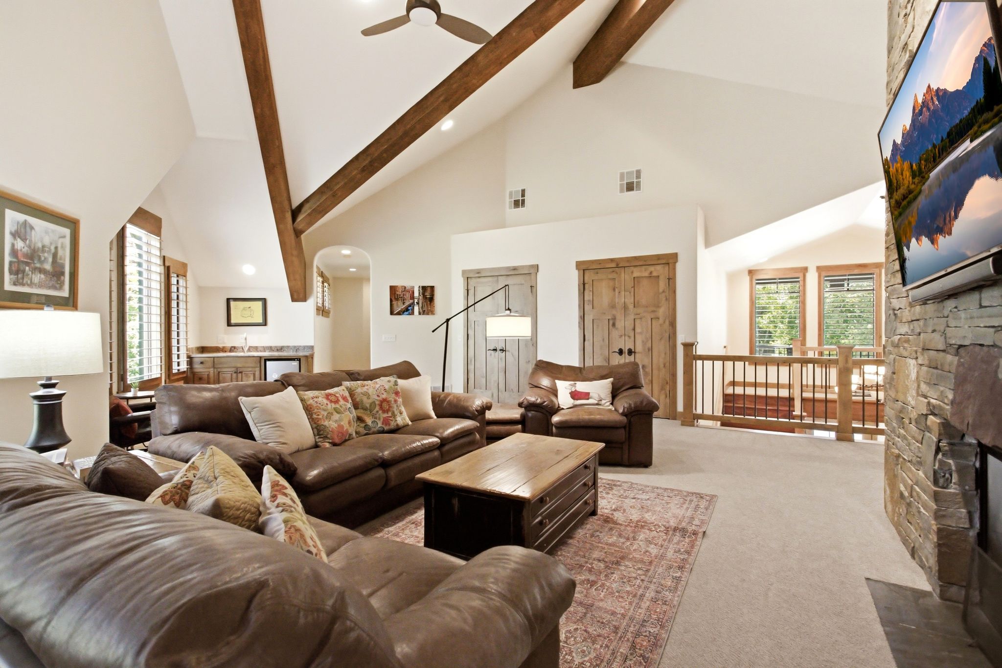 Vaulted ceilings with exposed wood beams, a stone-clad fireplace, and a built-in wet bar with cabinetry define this expansive living area